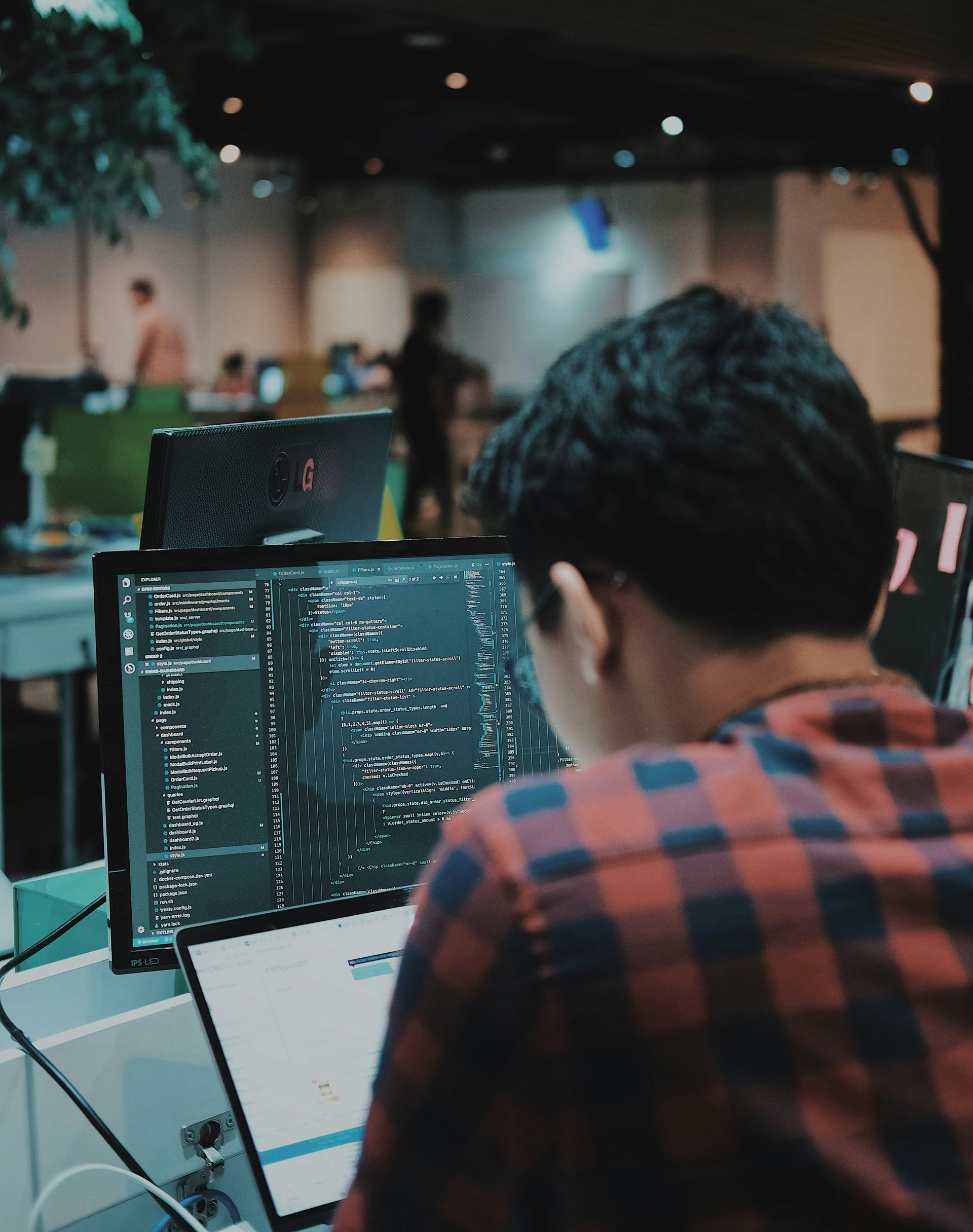 A person wearing a red plaid shirt works on code displayed on a desktop monitor and laptop in a modern office space.