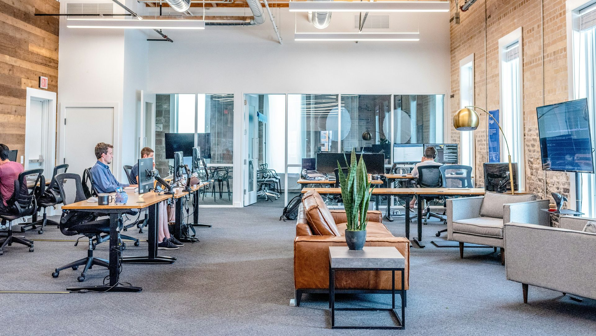 Modern office space with desks, computers, and employees. Exposed beams, brick walls, and open floor plan.