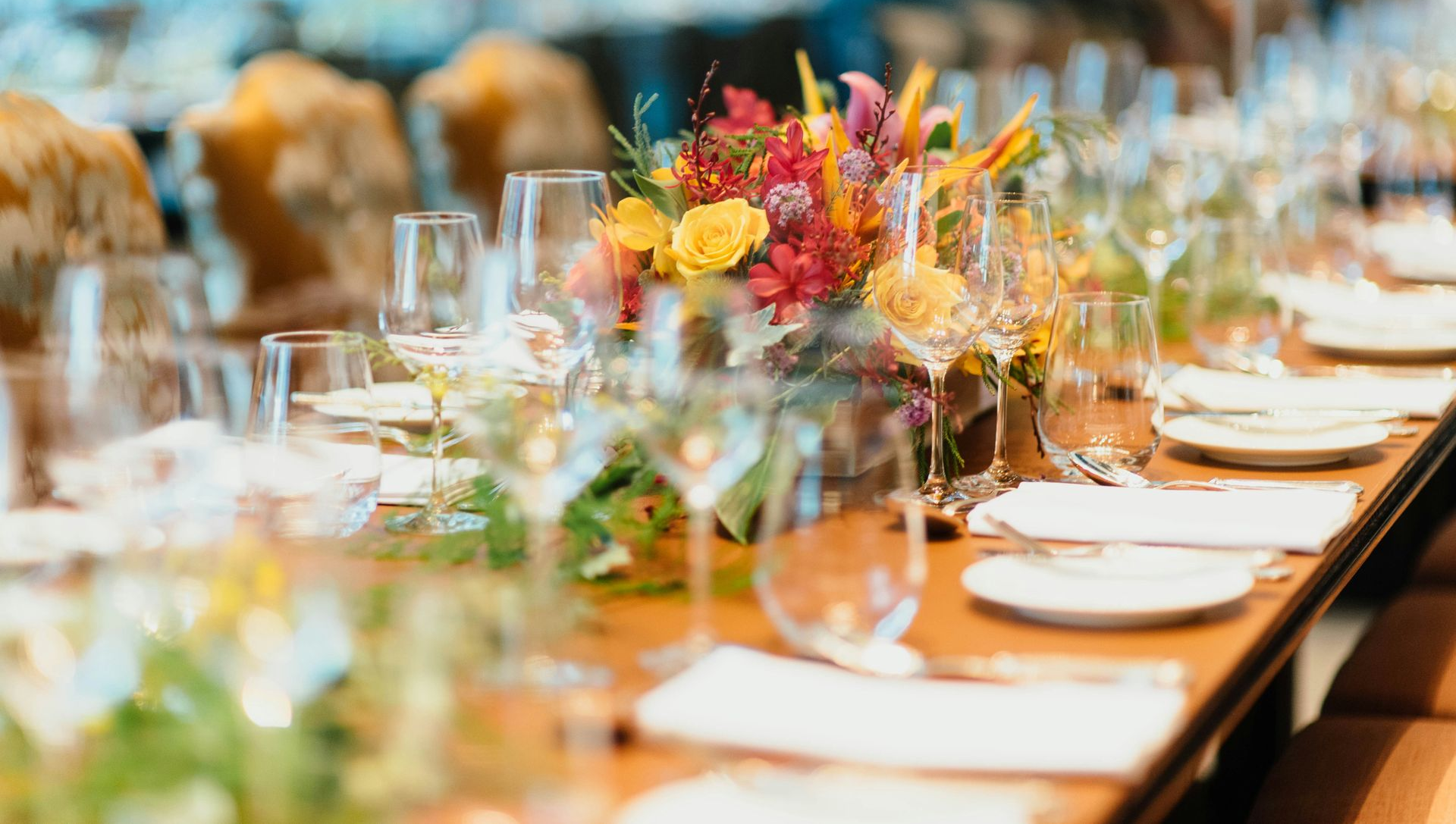A long dining table set for an event with glassware, white plates, and a vibrant floral centerpiece of yellow and red.