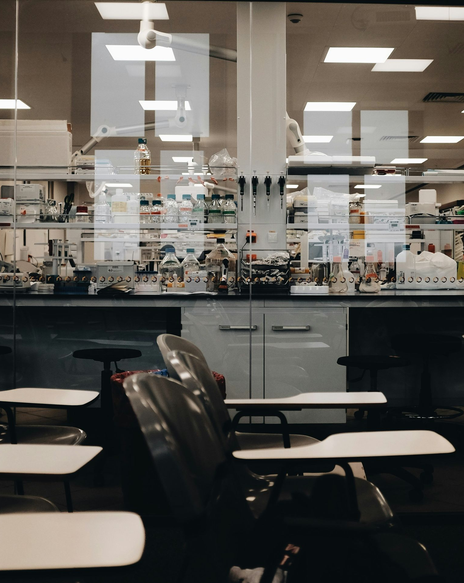 Empty classroom or lab with rows of desks and chairs, seen through a glass wall under bright ceiling lights