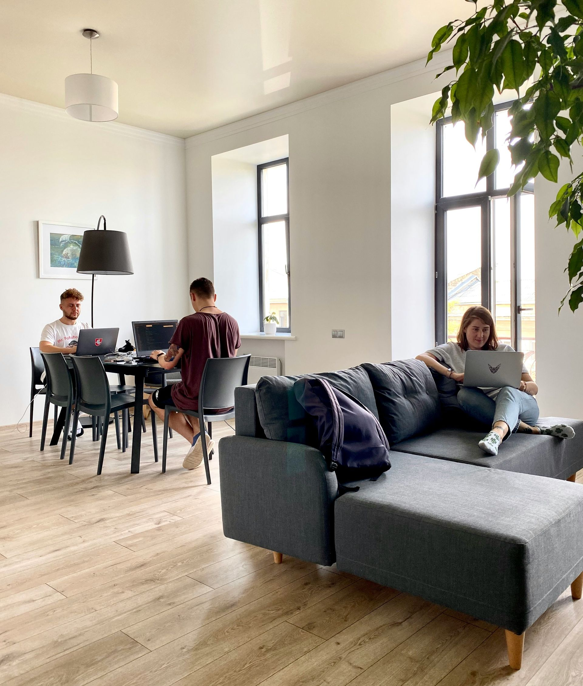 Bright living room with people working at a dining table and a person relaxing on a gray sofa