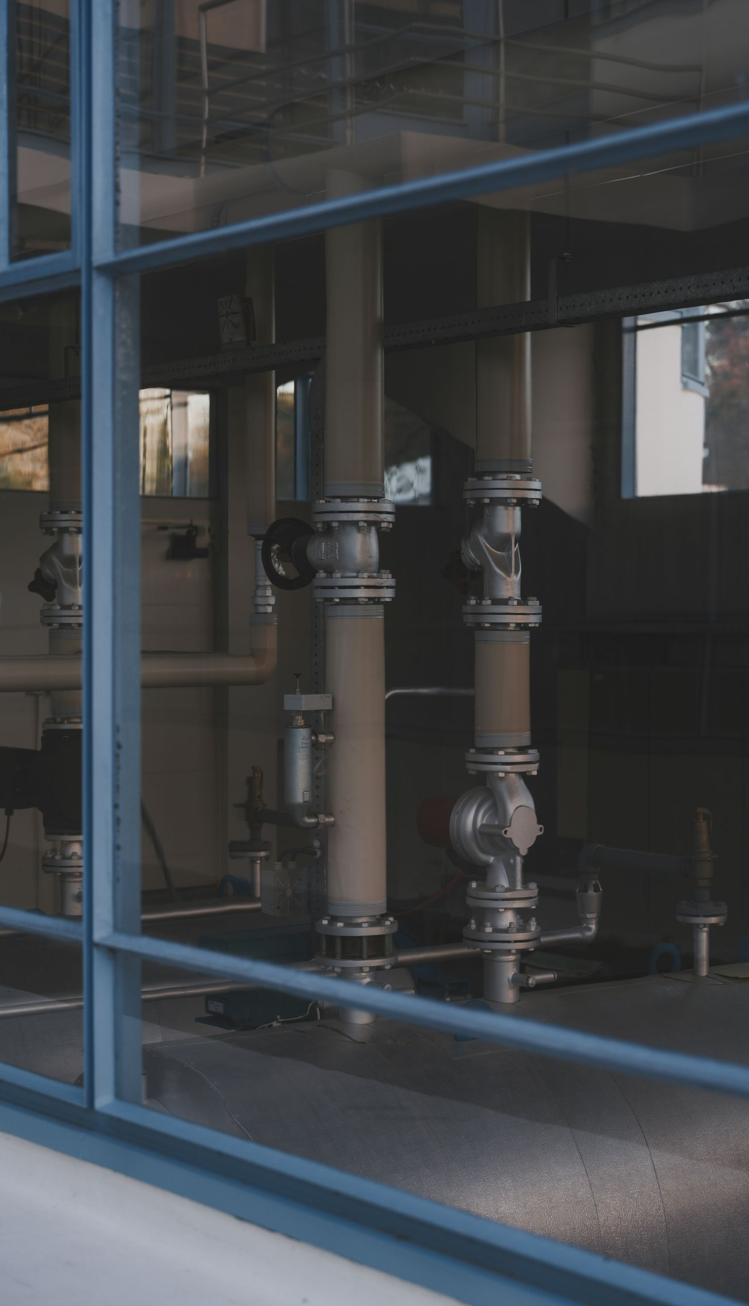 Vertical pipes with metallic valves seen through a glass window inside an industrial or mechanical facility.