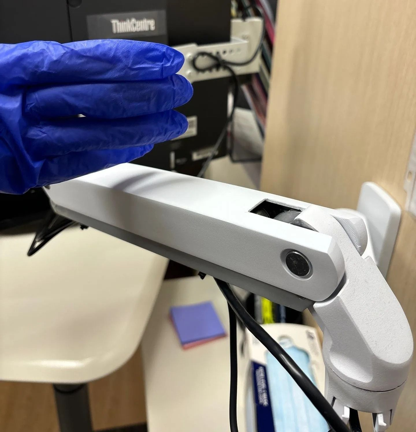 A hand in a blue glove over a white projector arm. A desk and computer are in the background.