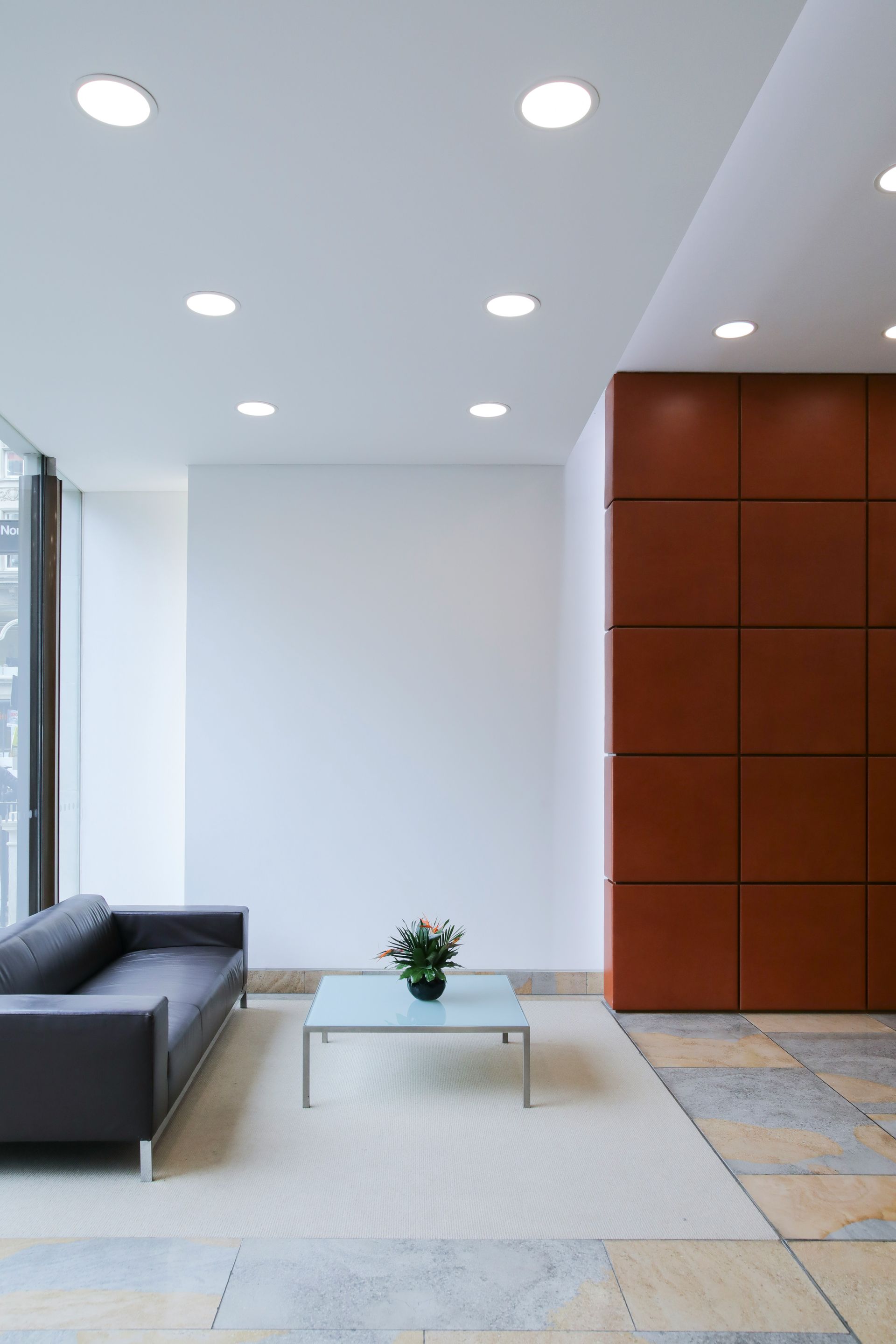 Modern office waiting area with dark sofa, glass table, and brown wall panels.