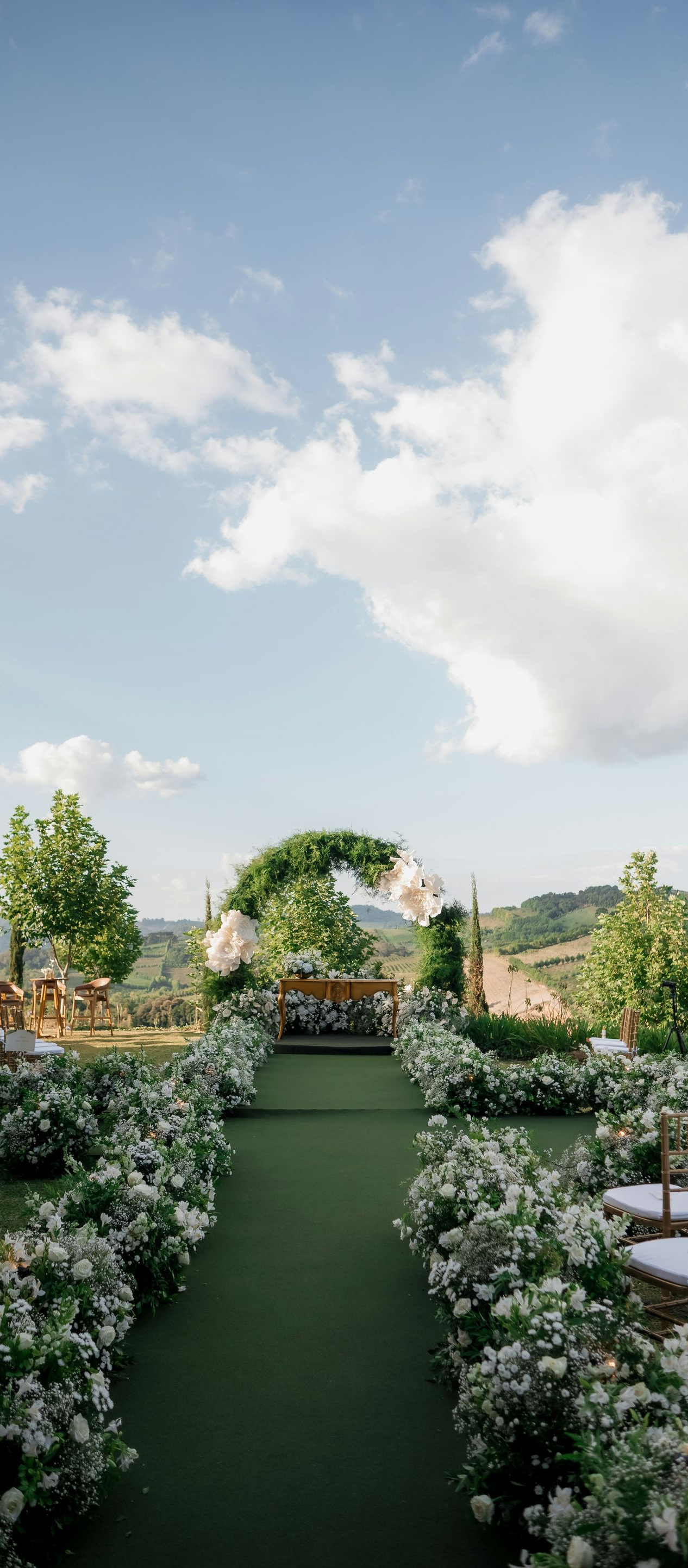 An outdoor wedding ceremony aisle lined with white flowers leads toward a circular altar against a scenic landscape.