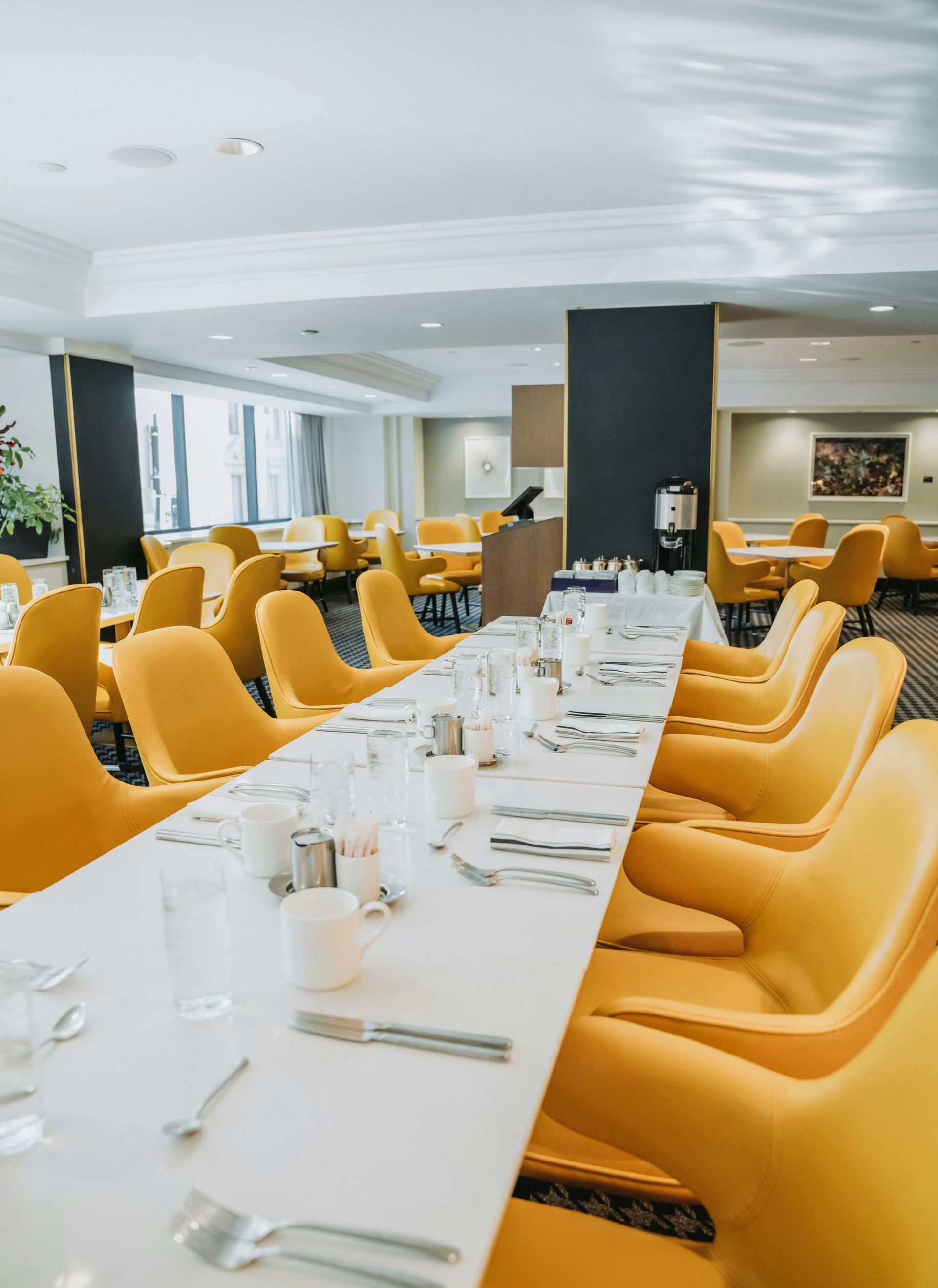 A long, empty dining table with place settings, flanked by rows of vibrant yellow modern chairs in a bright restaurant.