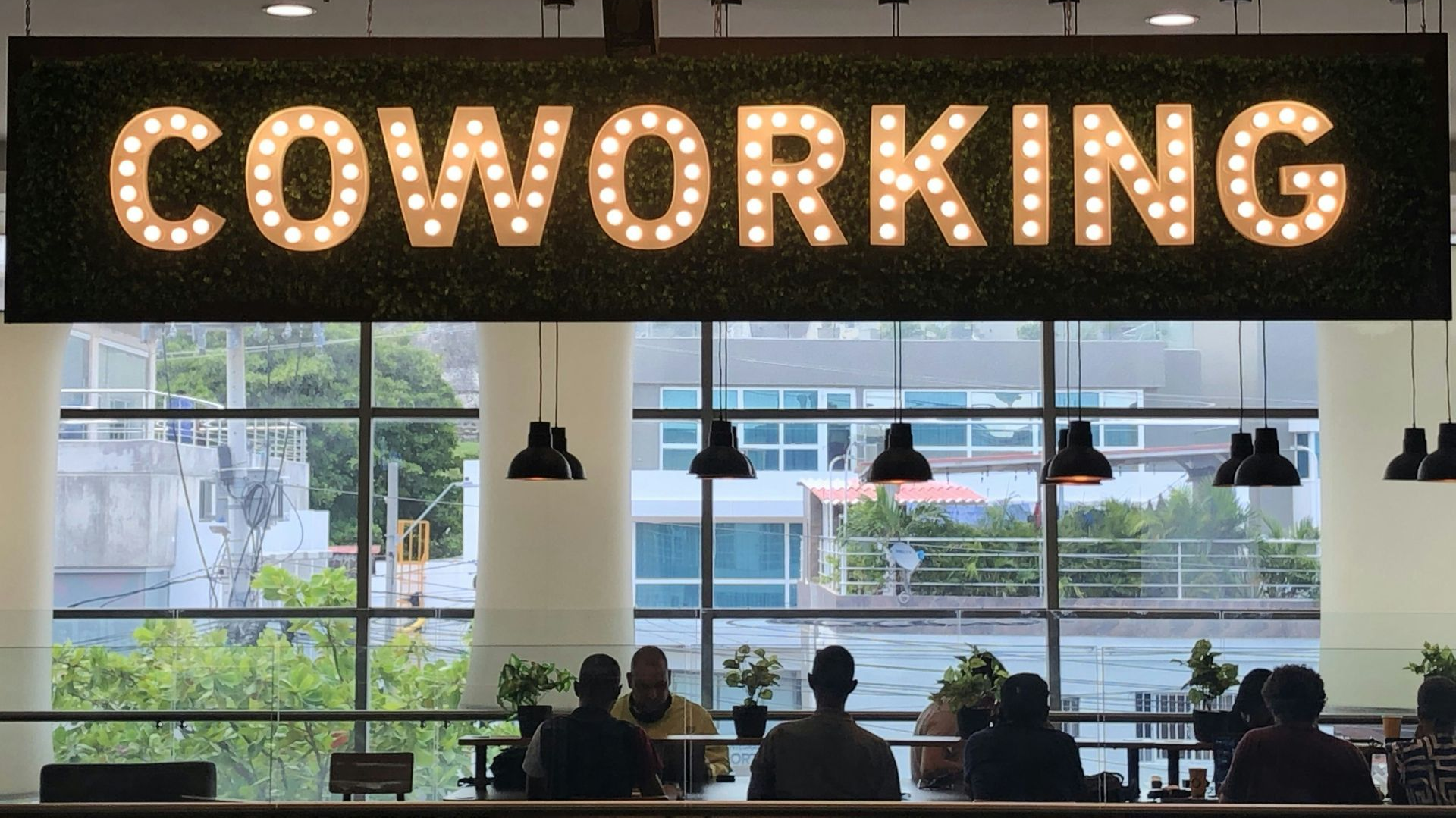 Bright “COWORKING” sign above people seated inside a modern workspace with large windows.