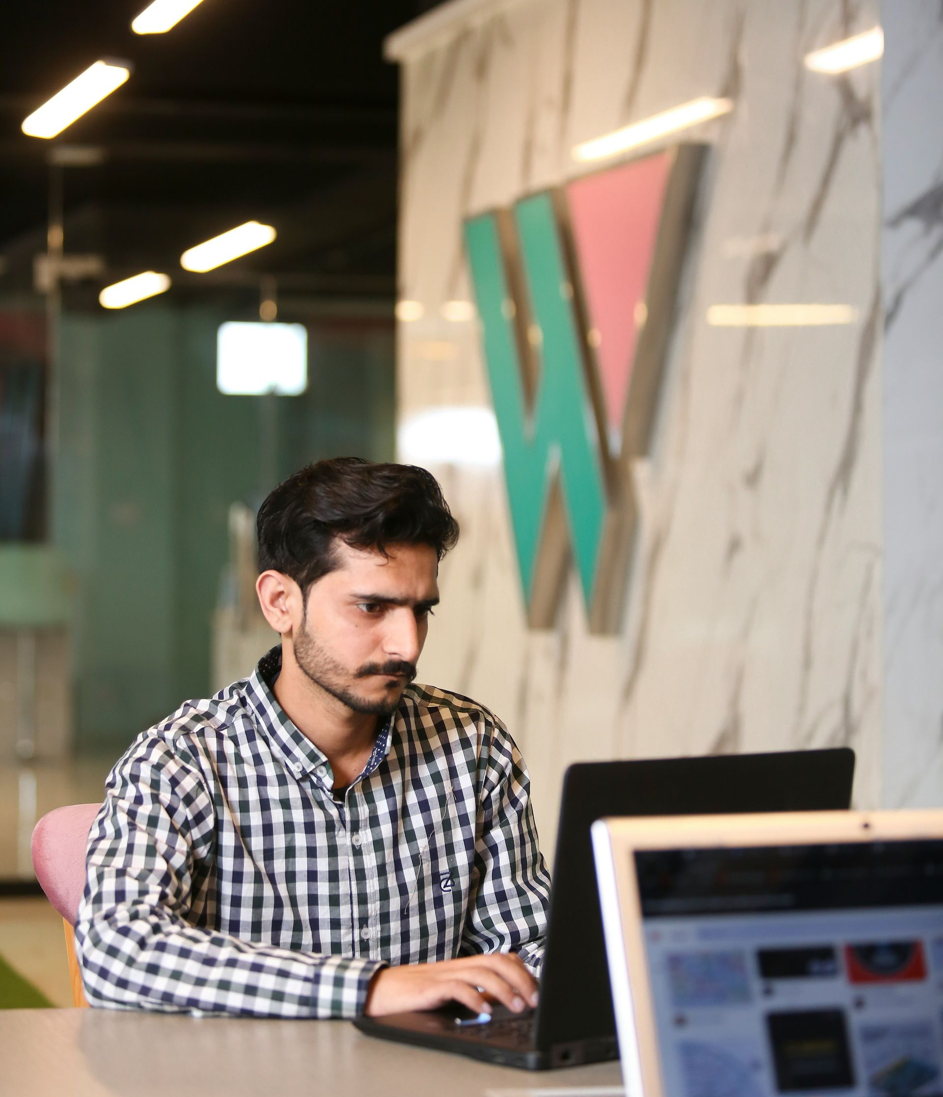 Man working on a laptop at a desk in a modern office with a large wall logo in the background