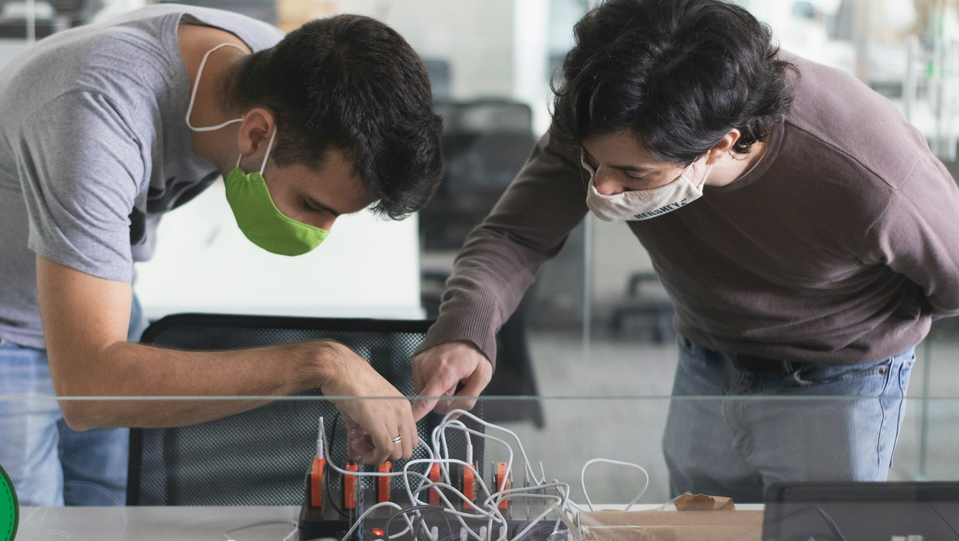 Two people lean over a tabletop electronics project, inspecting wires and components in a workshop.
