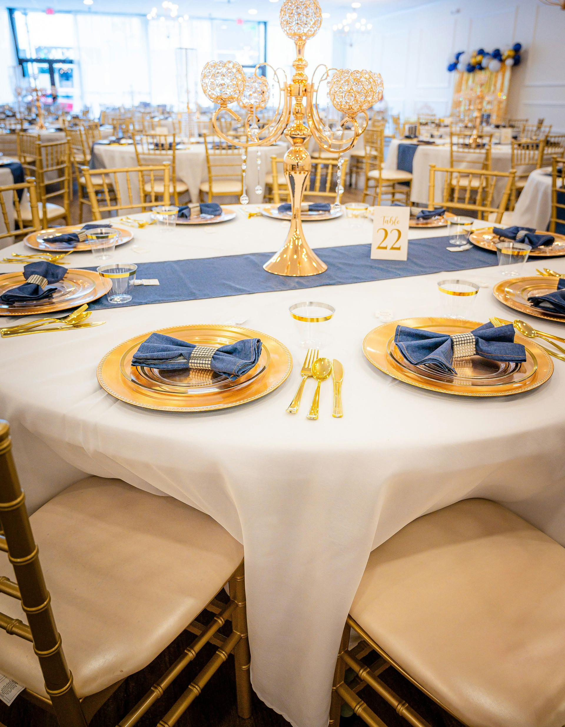 A round event table set with white linens, gold-rimmed plates, blue napkins, and a gold candelabra centerpieces.