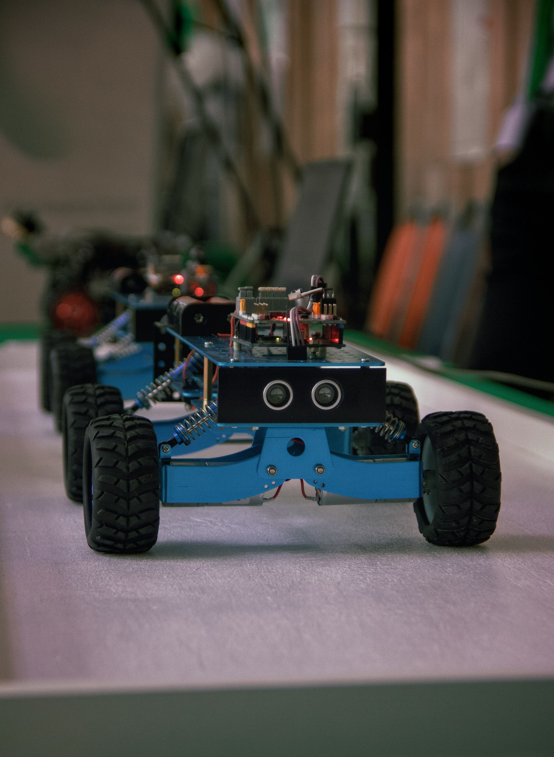 Blue wheeled robot car with exposed electronics on a tabletop, seen from the front.