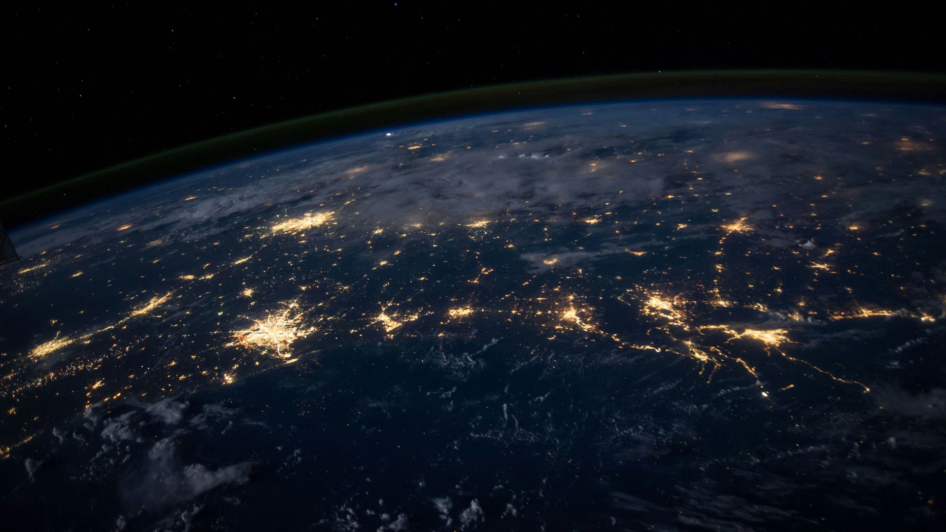 A satellite view of Earth at night, showing bright city lights glowing against the dark surface and the planet's horizon.