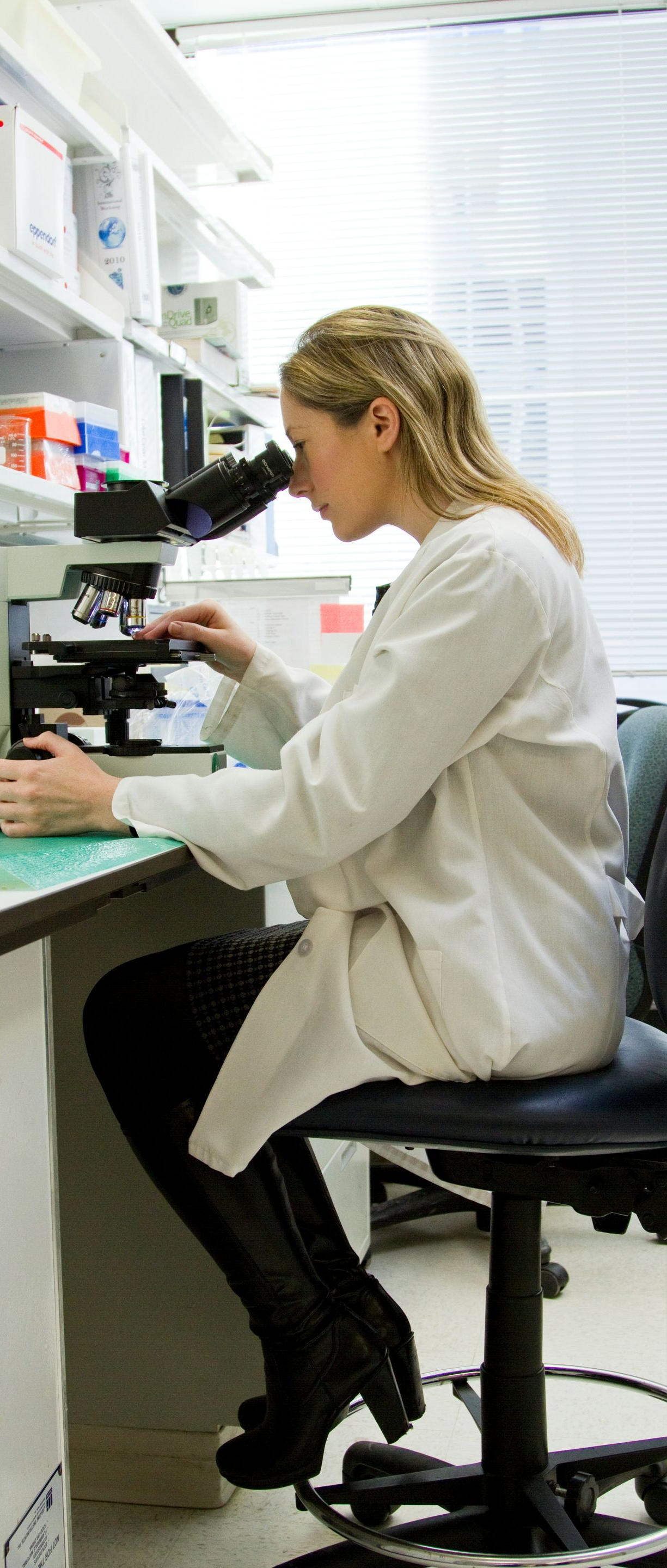 Scientist using a microscope at a lab bench with shelves of supplies behind her
