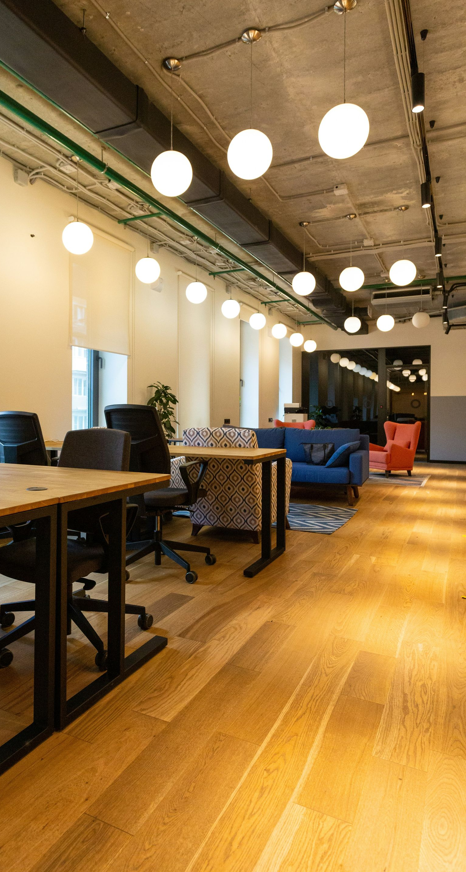 Open office lounge with long wooden tables, blue chairs, and hanging globe lights on a wood floor.