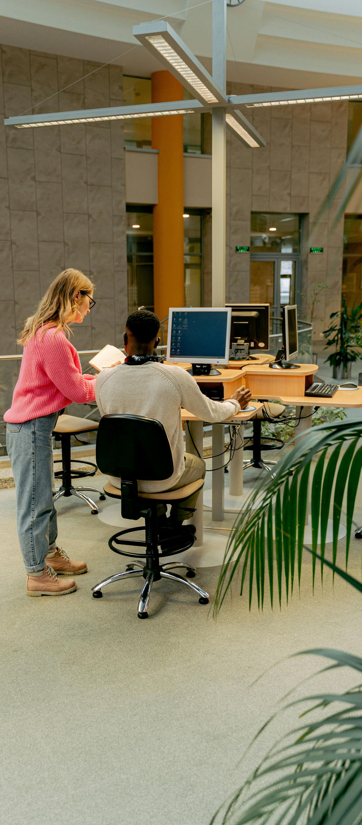 Two people at computers in a modern office lobby, with tables, columns, and plants nearby.