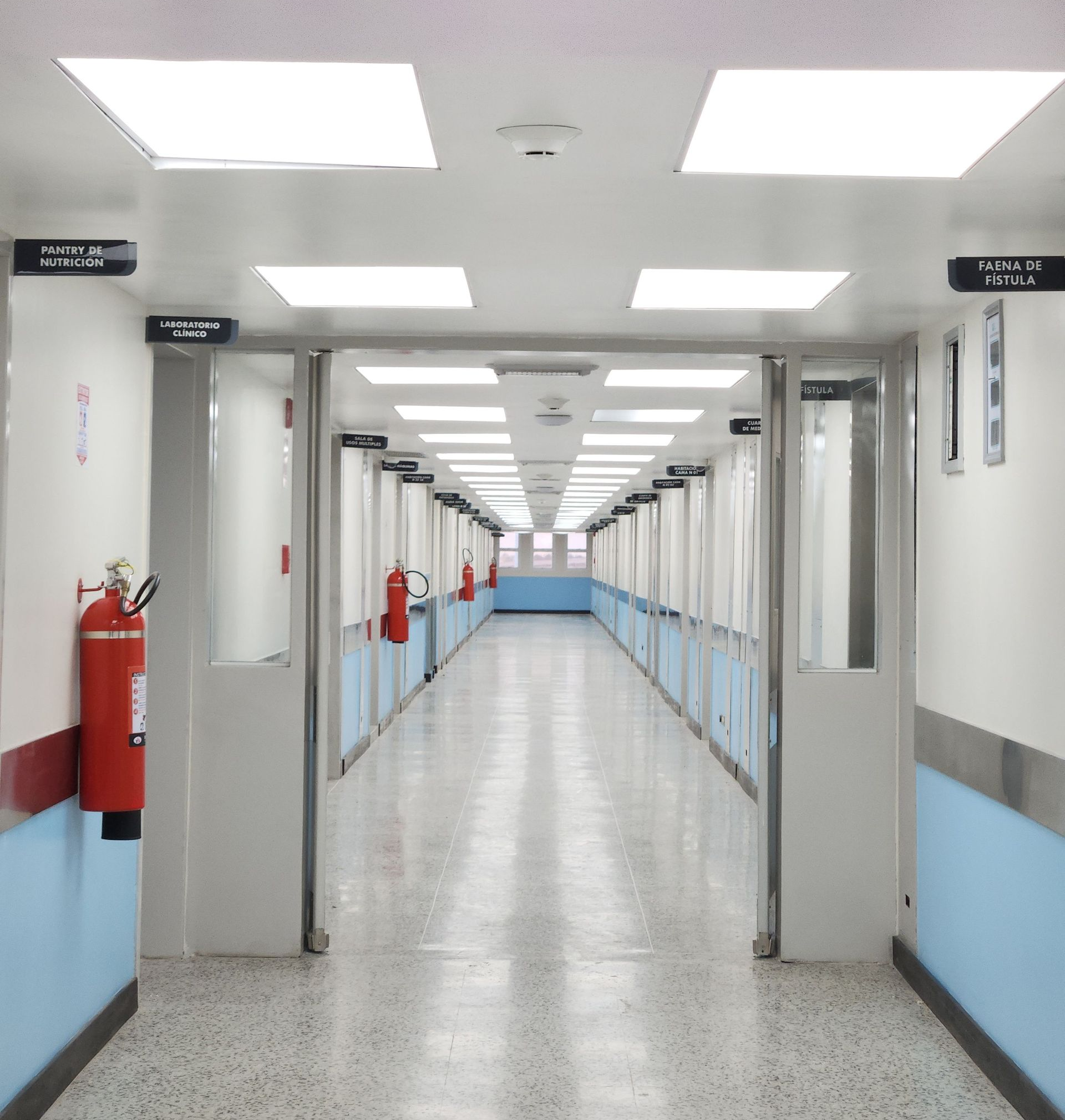 Empty hospital corridor with bright ceiling lights, white walls, blue lower trim, and fire extinguishers