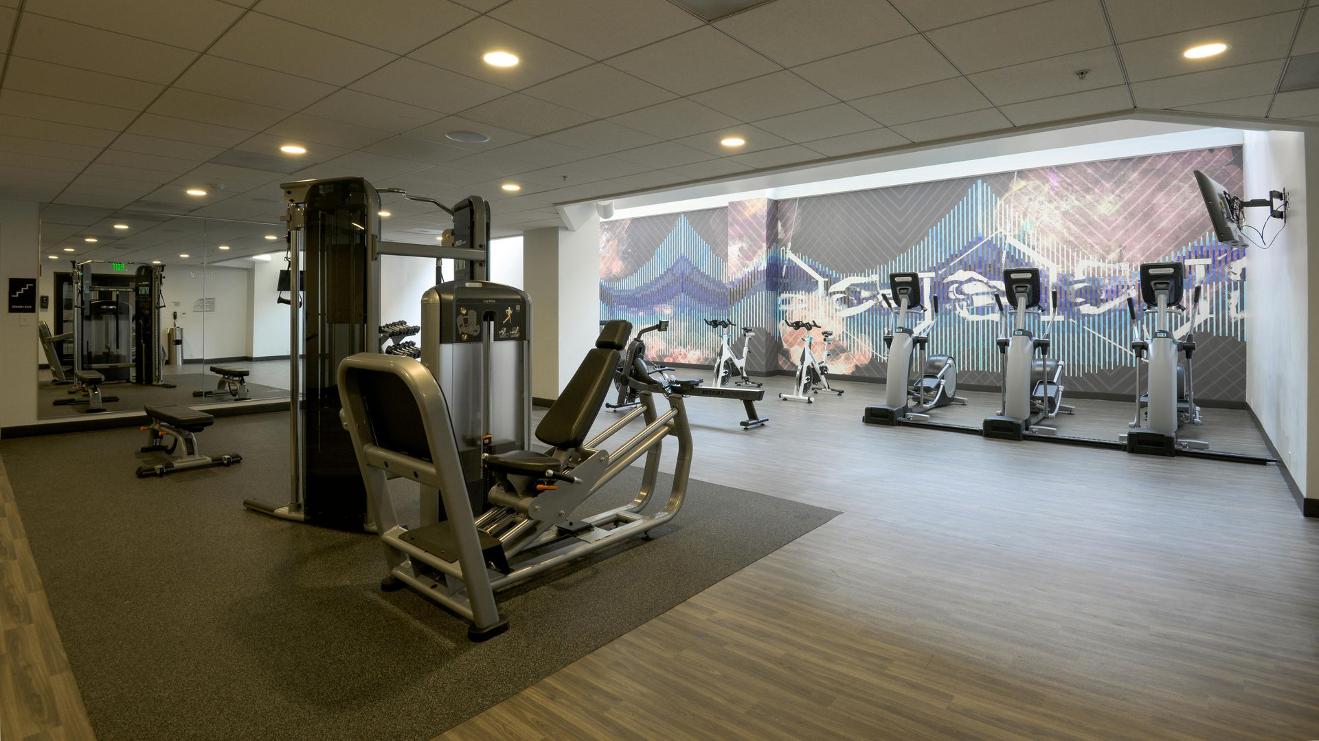 A gym interior with exercise machines, including weight equipment and treadmills, set against a city-themed mural wall.