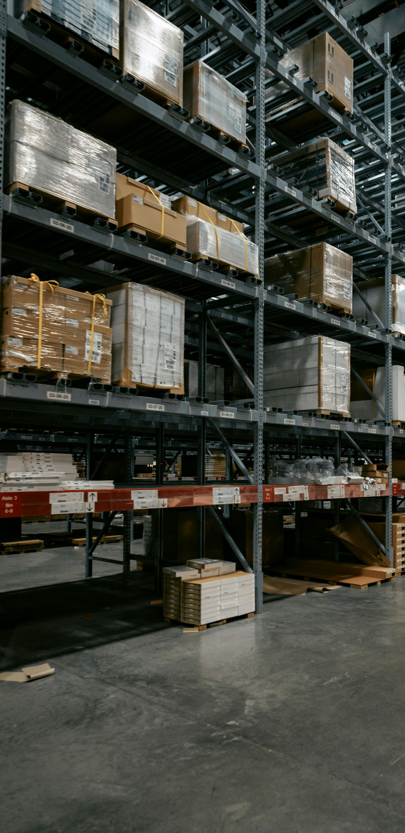 Tall metal industrial shelving units stocked with cardboard boxes and palletized goods in a warehouse setting.