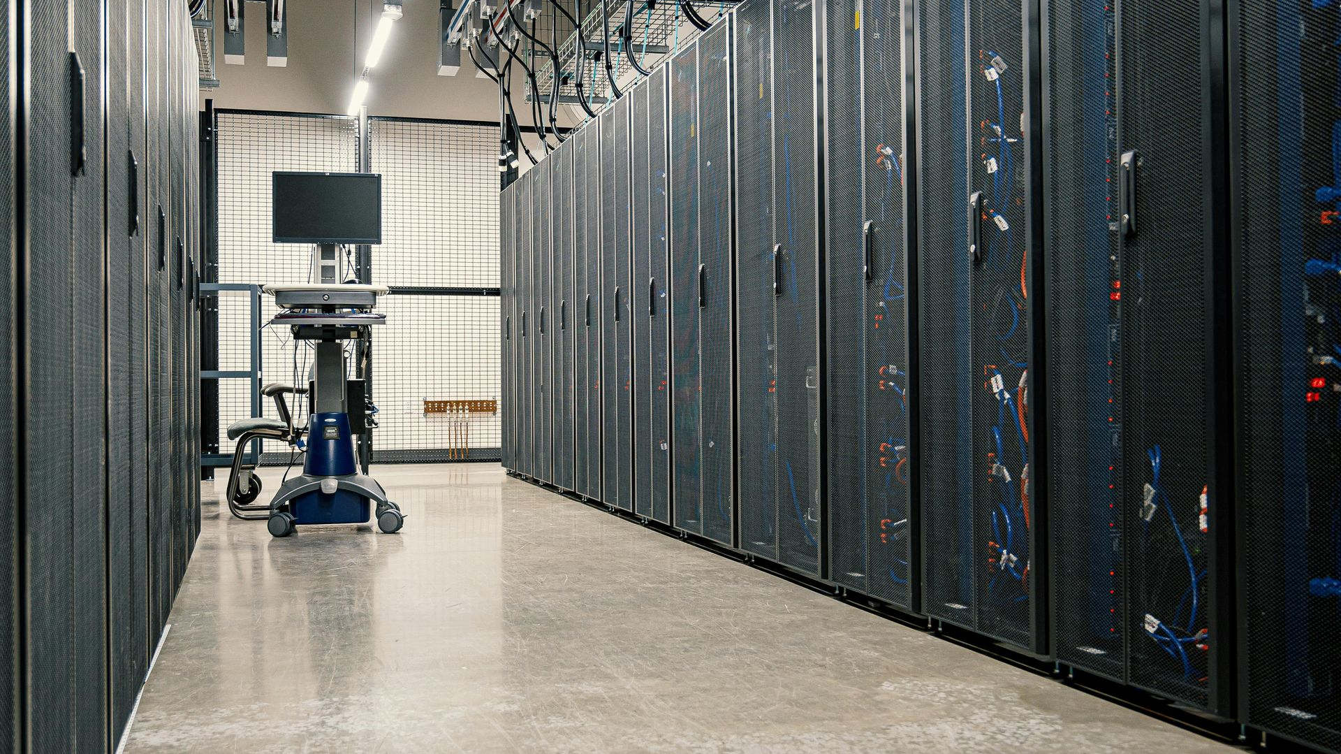 A server room with rows of tall black server cabinets on a polished floor and a mobile monitor cart in the background.