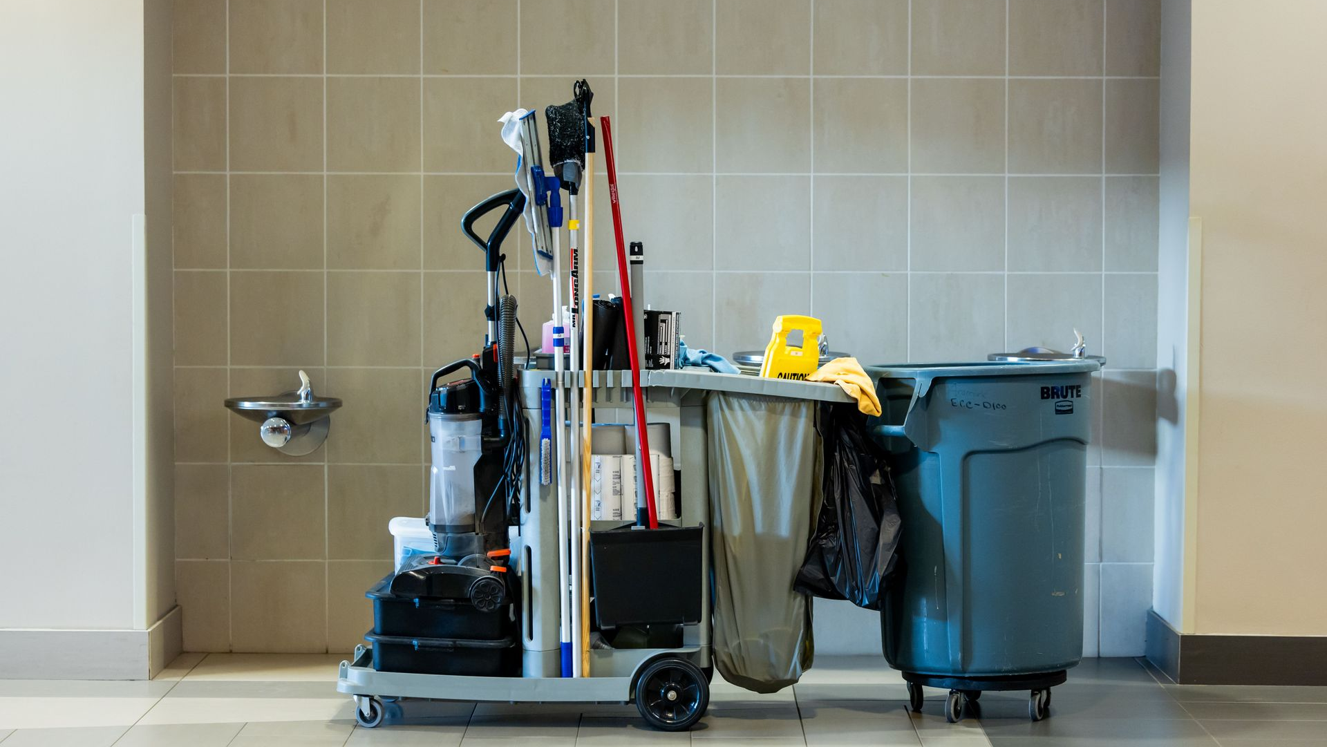 Janitorial cart with cleaning supplies and mop beside trash bins in a tiled hallway