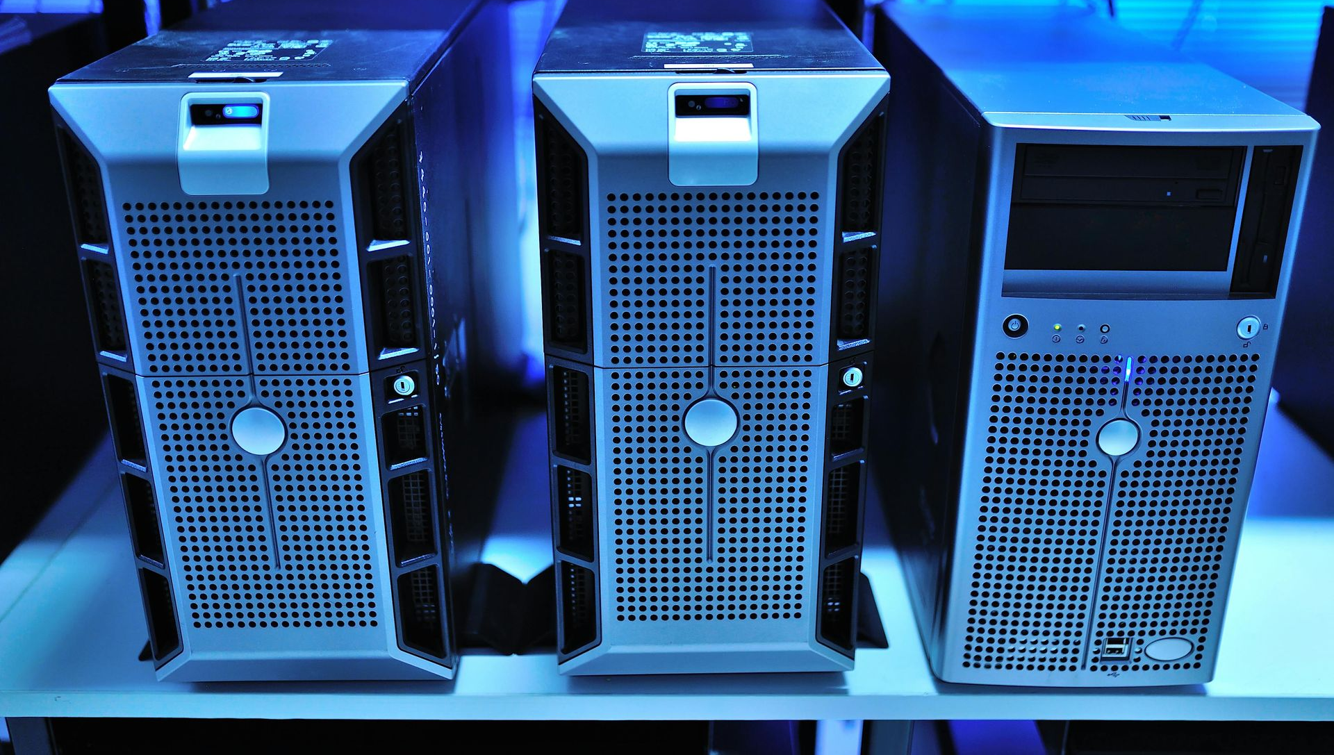 Three Dell PowerEdge tower servers sitting side-by-side on a shelf under blue lighting.