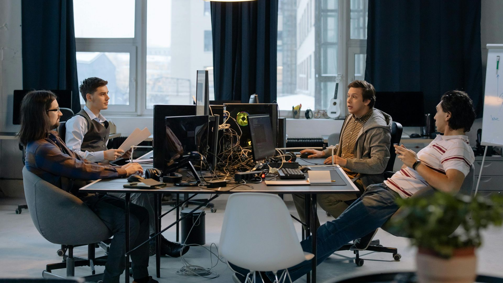 Four people in a modern office meeting around a table with laptops and monitors.
