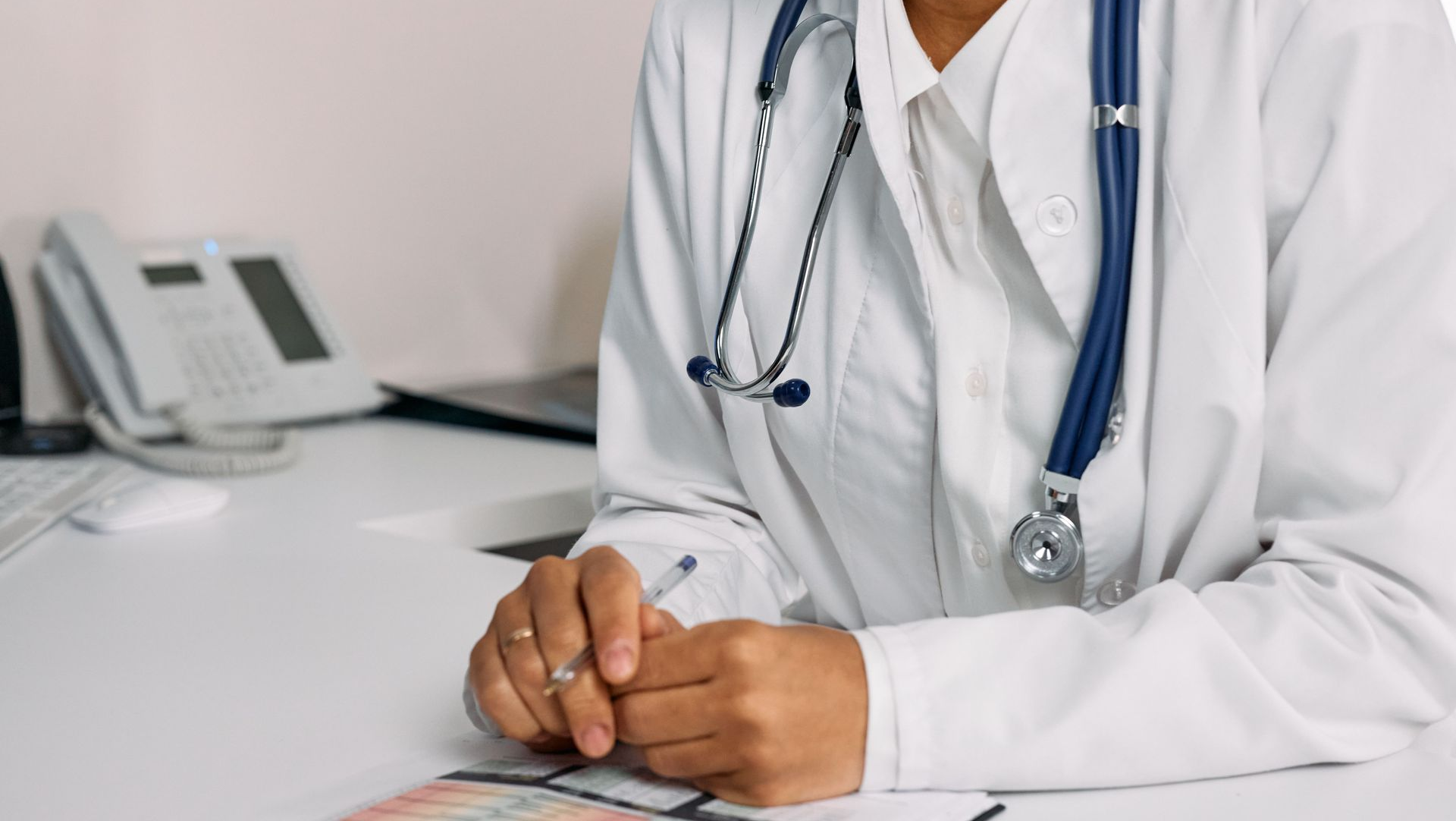 Doctor in a white coat with a stethoscope, seated at a desk with hands clasped in a clinic office.