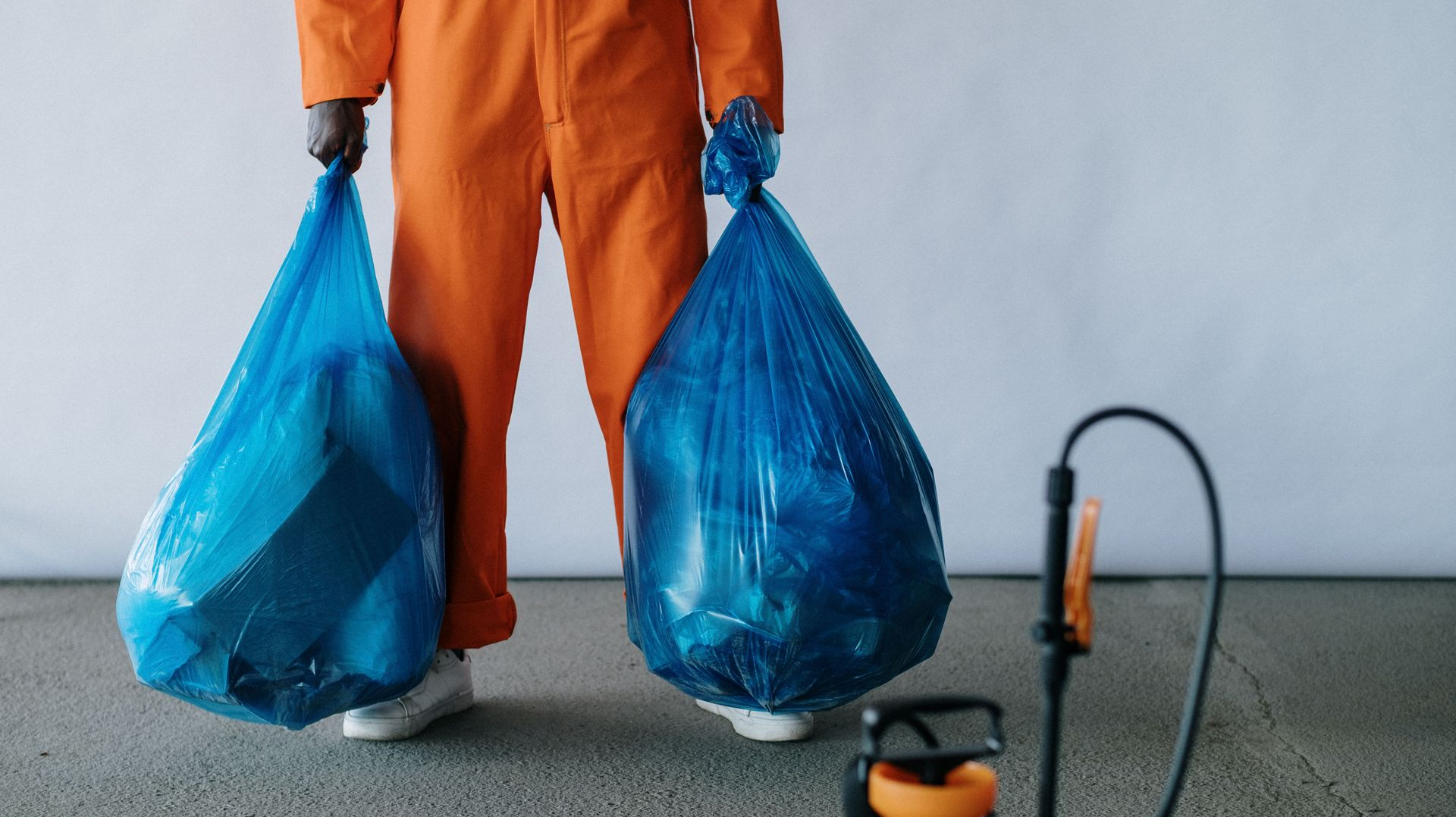 Person in orange coveralls holding two blue garbage bags next to a vacuum cleaner on a floor.