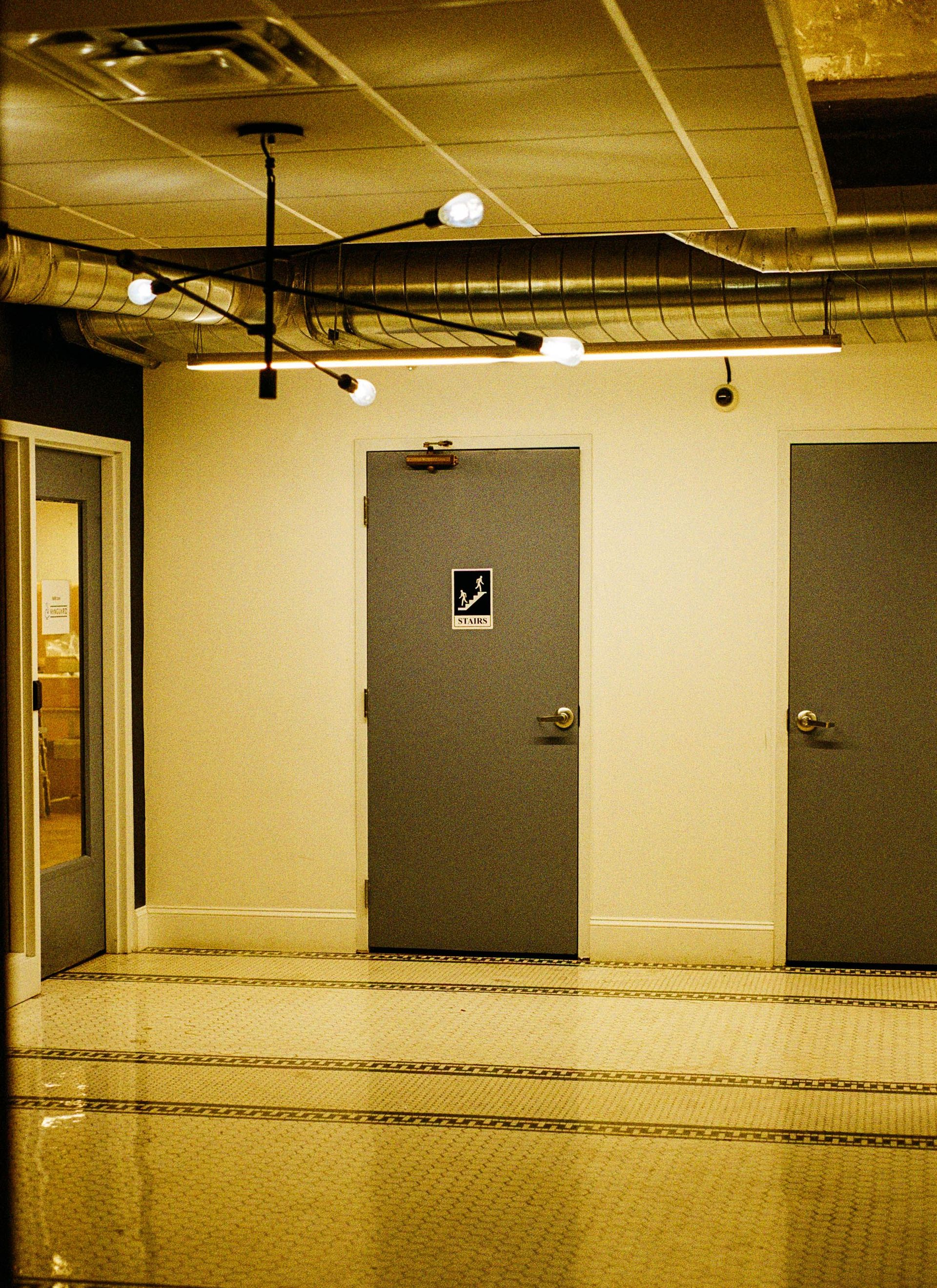 Empty hallway with gray doors, exposed ceiling pipes, and beige walls under warm lighting