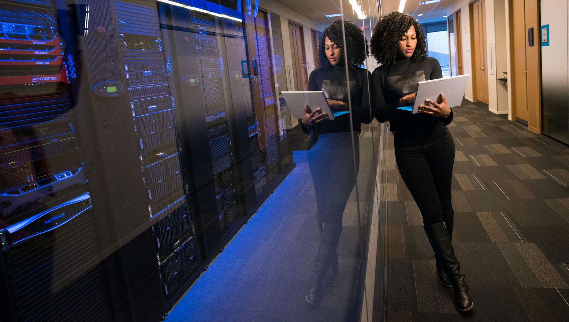A person stands in a server room hallway, holding a tablet and leaning against a glass wall reflecting computer racks.