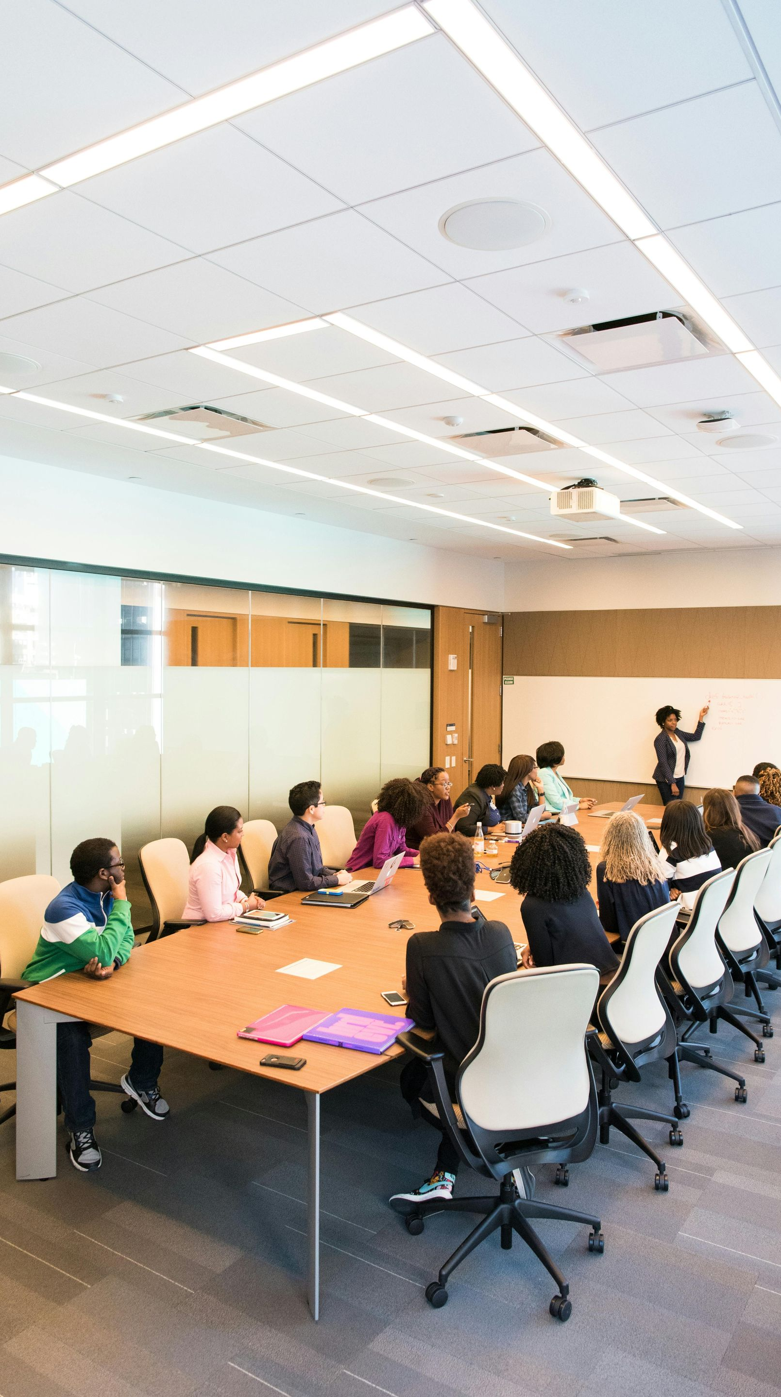 People seated around a table in a modern conference room, a person at the whiteboard giving a presentation.