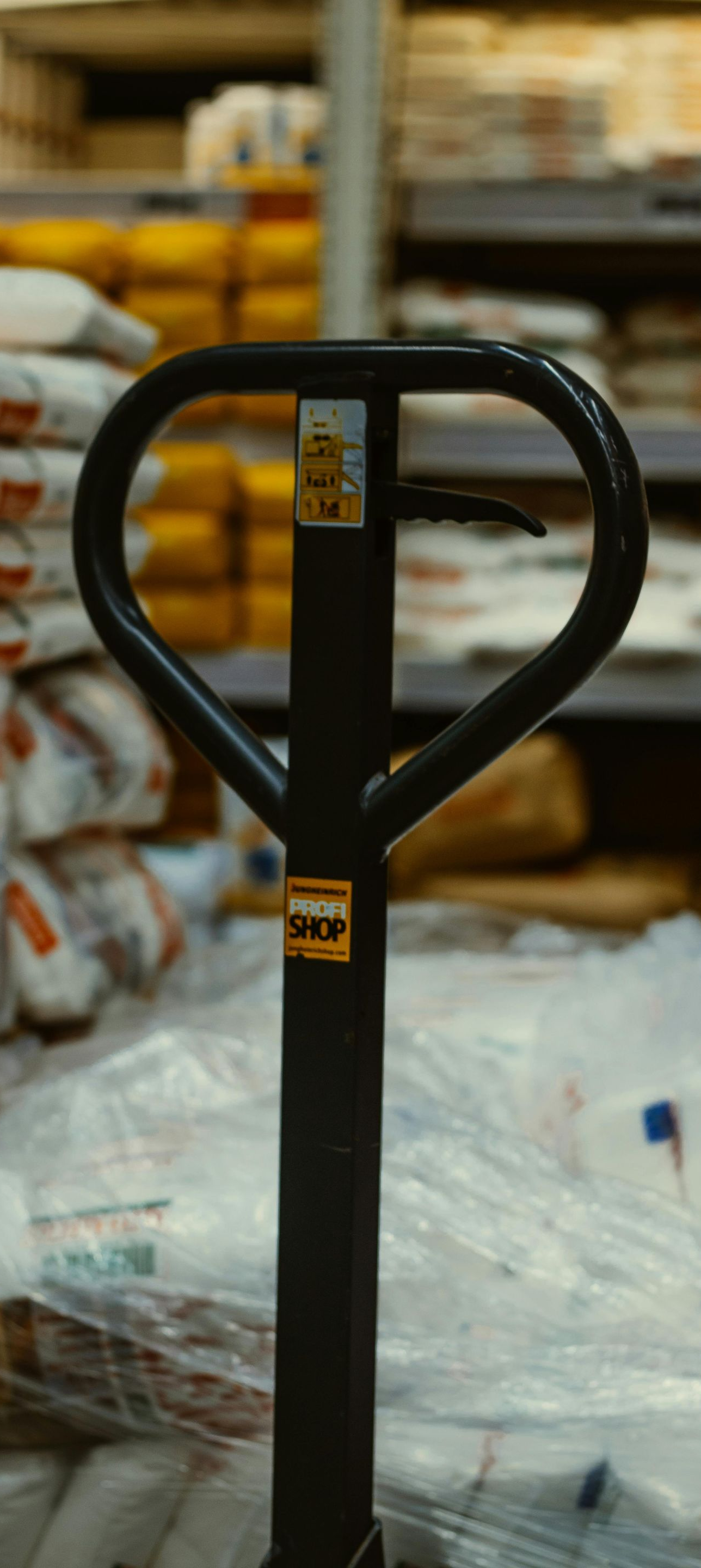 A black manual pallet jack handle in a warehouse setting, with shelves of stacked goods blurred in the background.