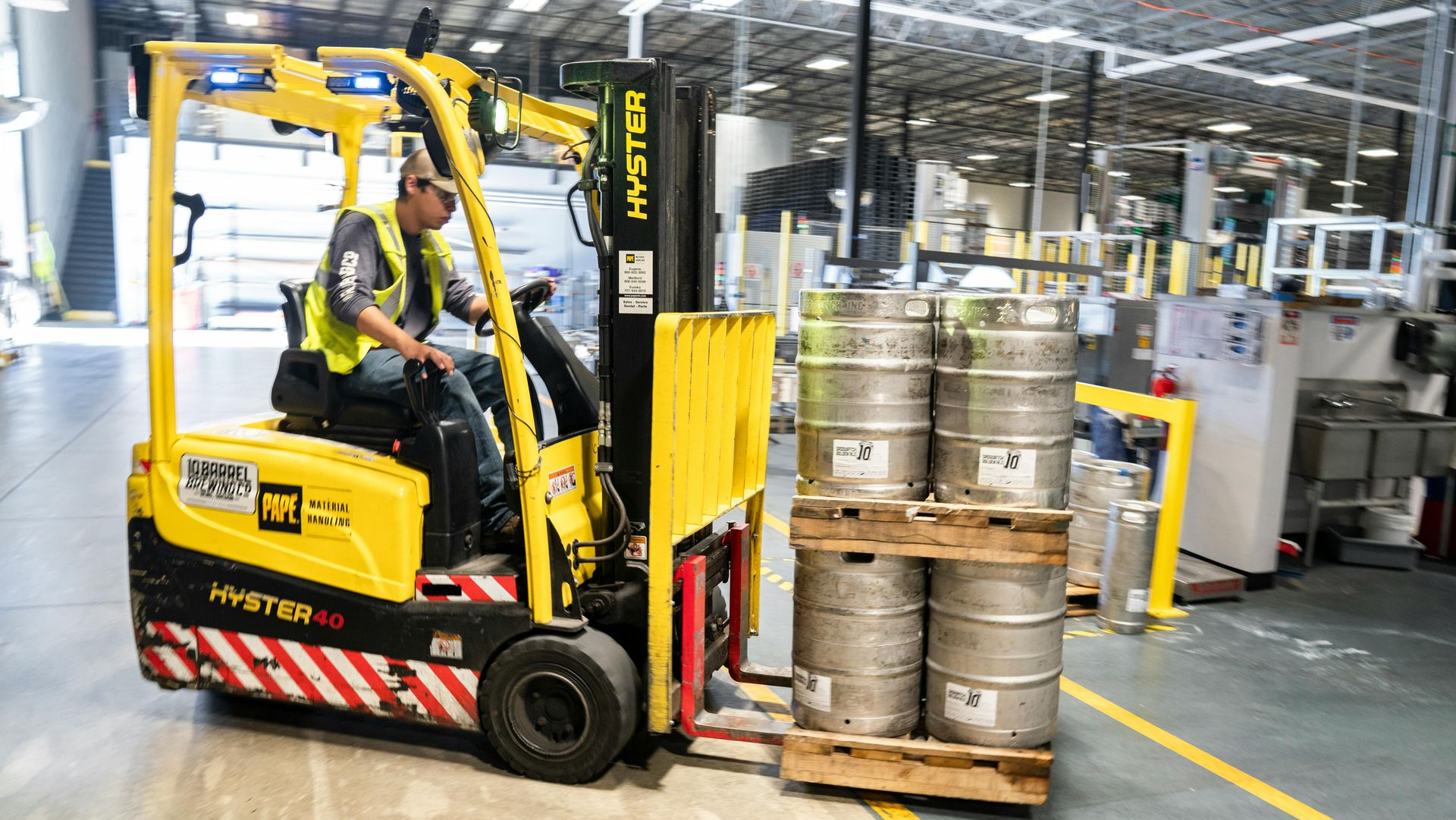 Forklift operator in yellow vest transports kegs in a warehouse.