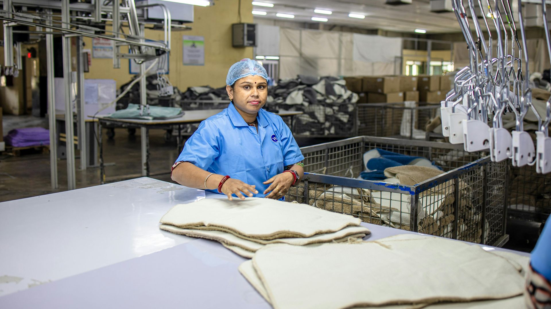 A worker in a blue uniform and hairnet works at a table in a garment factory, preparing stacks of white fabric.