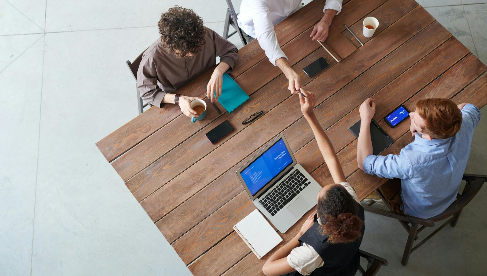 Team meeting around a wooden table with laptops, coffee cups, and two people shaking hands over a smartphone.