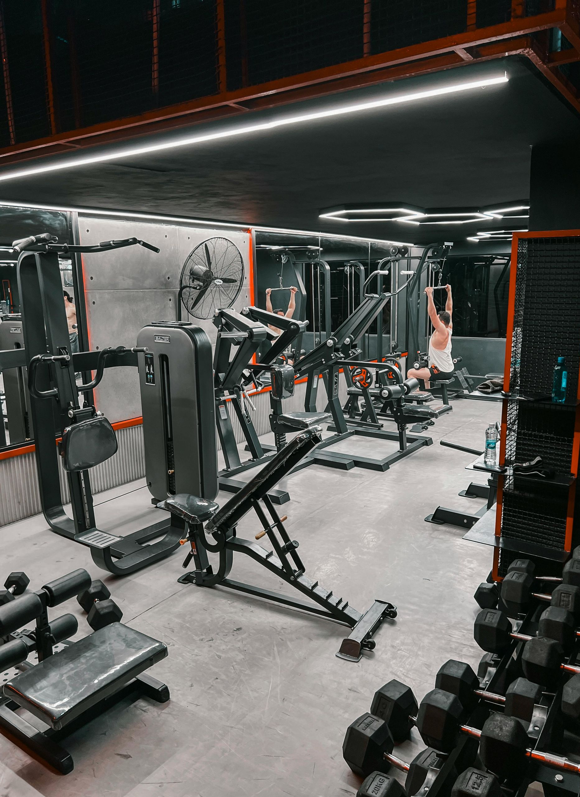 Modern gym with weightlifting equipment, dumbbell racks, and a person using a cable machine under bright neon lights.