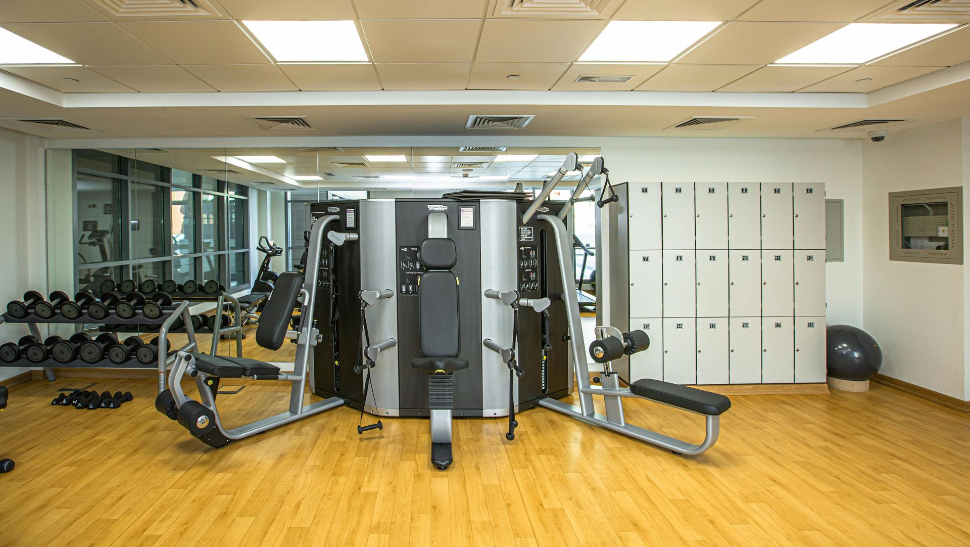 A well-lit, empty gym interior featuring a multi-station exercise machine, rows of lockers, and a dumbbell rack.