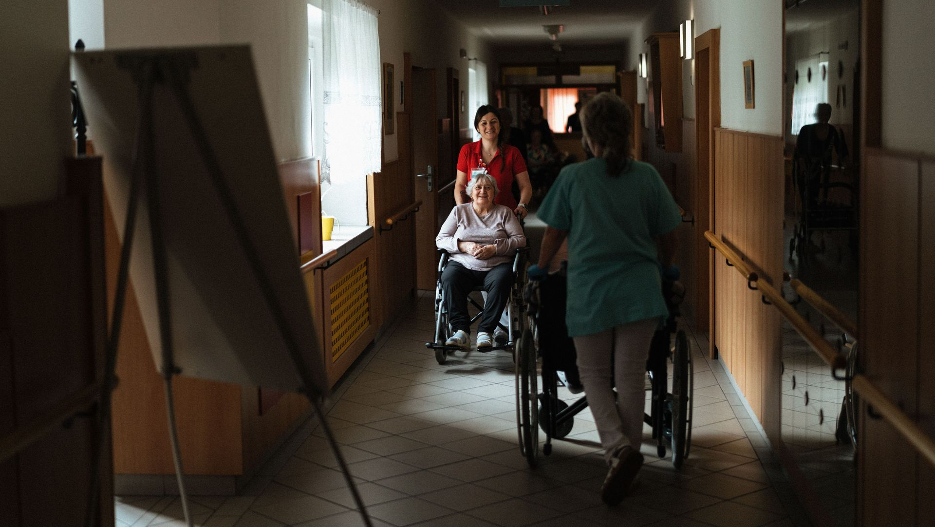 Two people in a dim hallway, one pushing a wheelchair toward the camera.