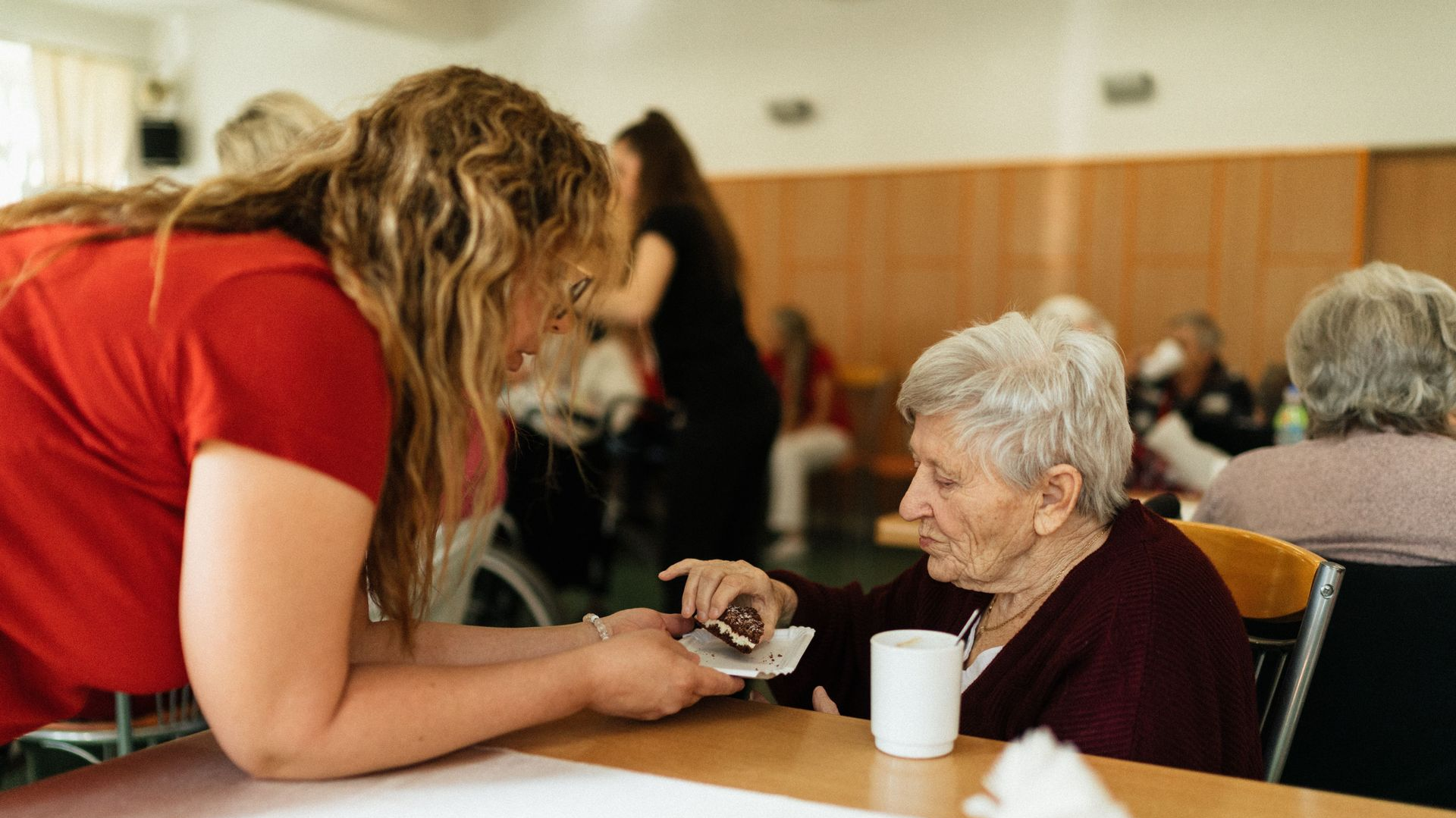 Caregiver leans over table to hand an elderly woman a cup in a warm community room