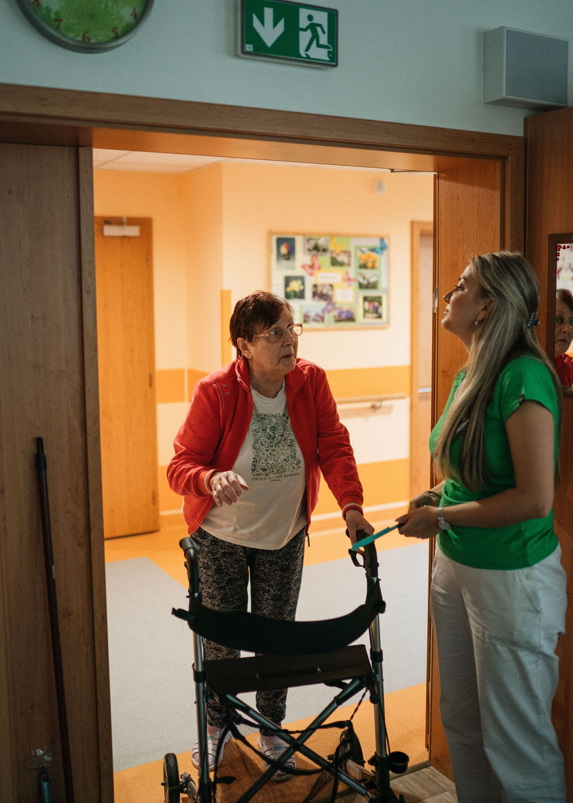 Two people talking beside a wheelchair in a hallway entrance with a green exit sign above.