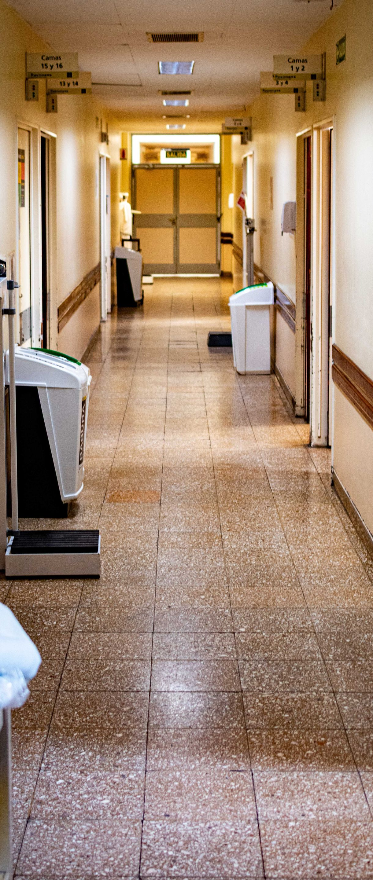 Long hospital corridor with beige walls, handrails, and trash bins under fluorescent lights