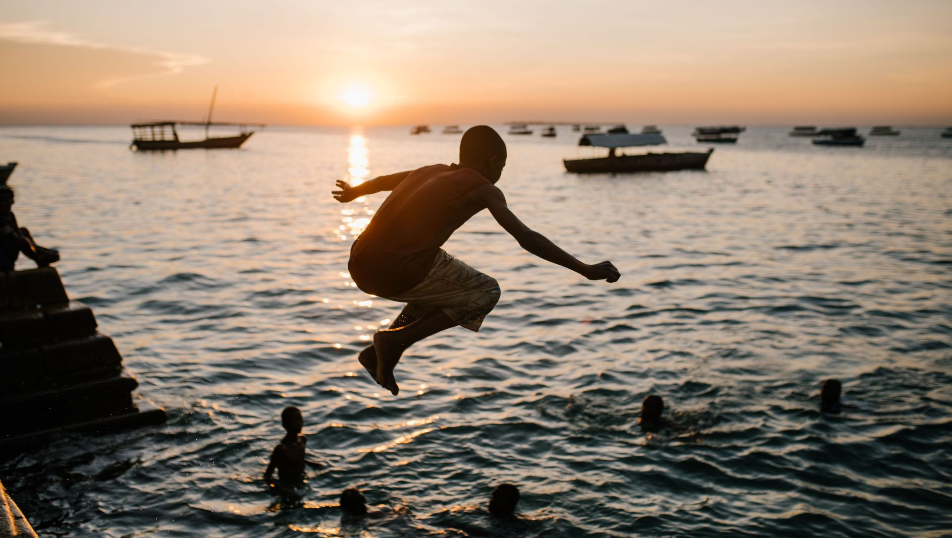 Silhouetted person jumping into the sea at sunset with boats in the background