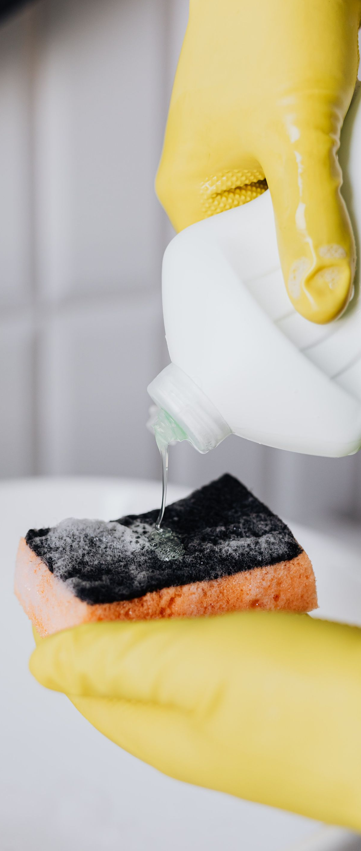 Yellow-gloved hands pouring liquid onto a black-and-orange sponge in a tiled sink area