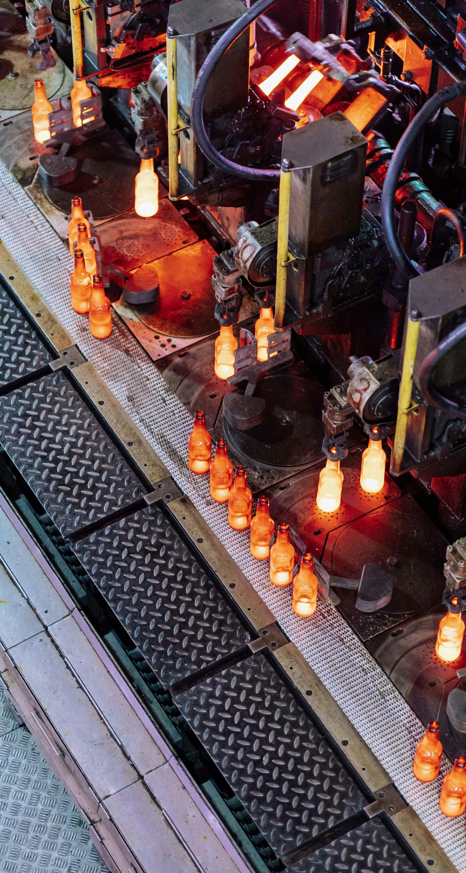 A high-angle view of a glass manufacturing line with glowing, molten glass bottles moving along a metal conveyor belt.