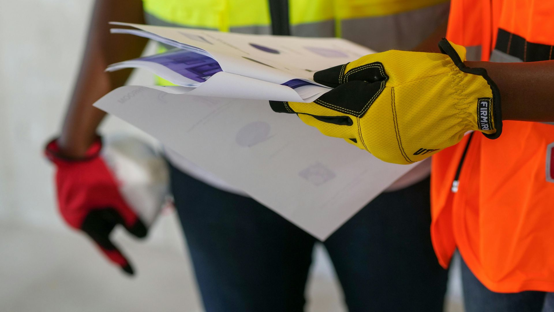 Person in safety vest and gloves holding papers with blue markings. Another person's red-gloved hand visible.