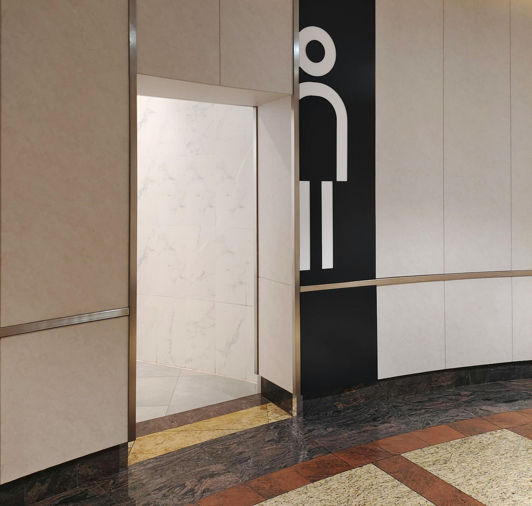 Entrance to a public restroom. Sign with a stylized person. Tan and black walls. Marble floors.