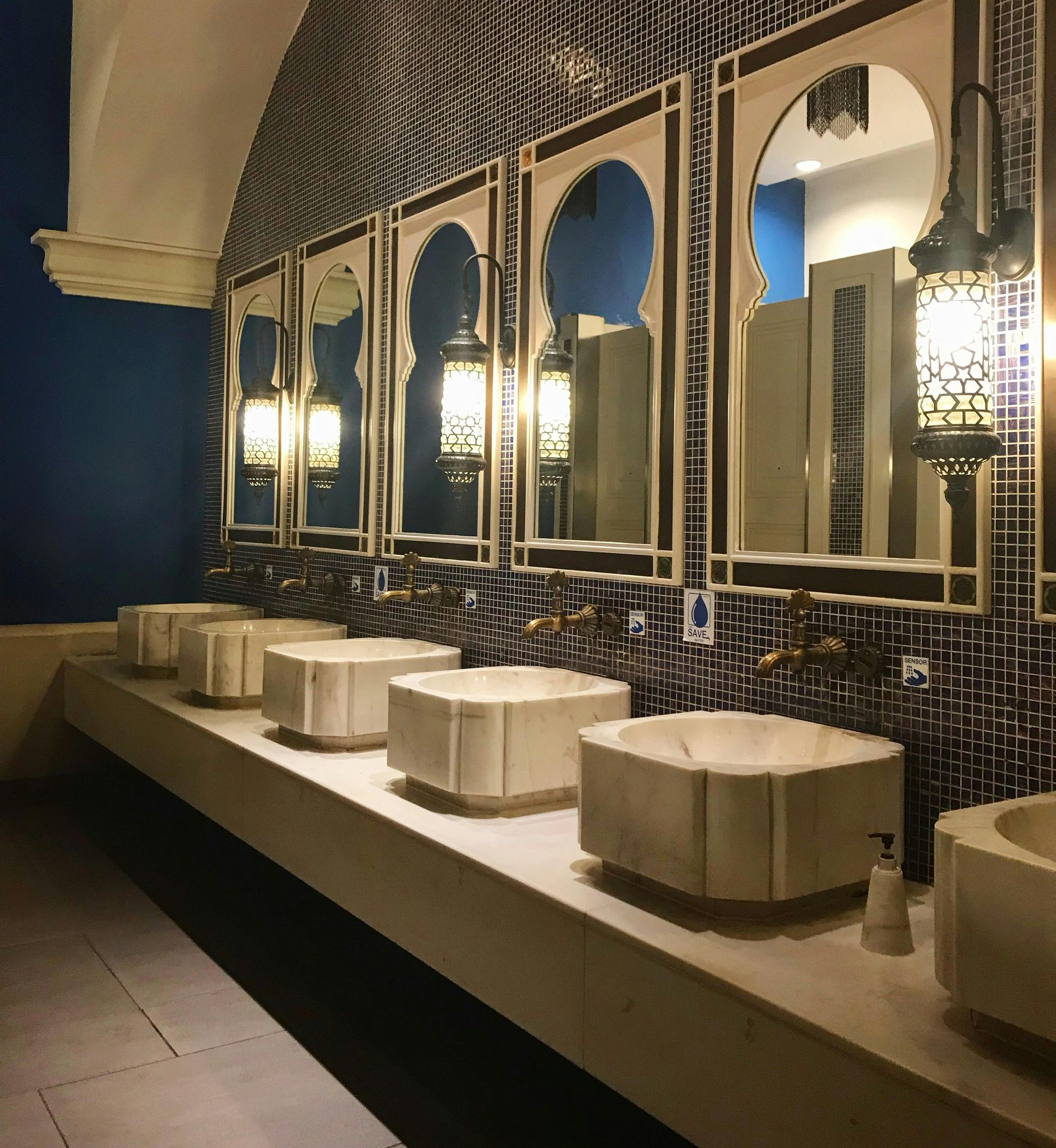 Public restroom with ornate mirrors, sinks, and Moroccan-style lighting, in a space with blue and white tiles.