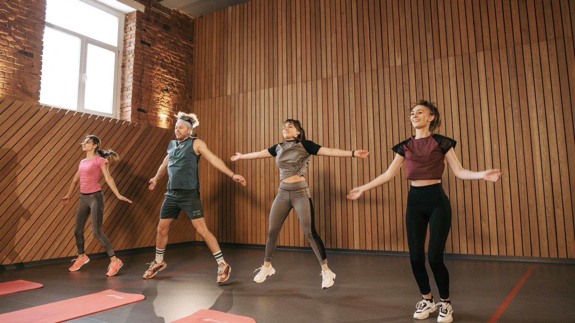 A group of people wearing athletic clothes jumping in a room with wood-paneled walls and yoga mats on the floor.