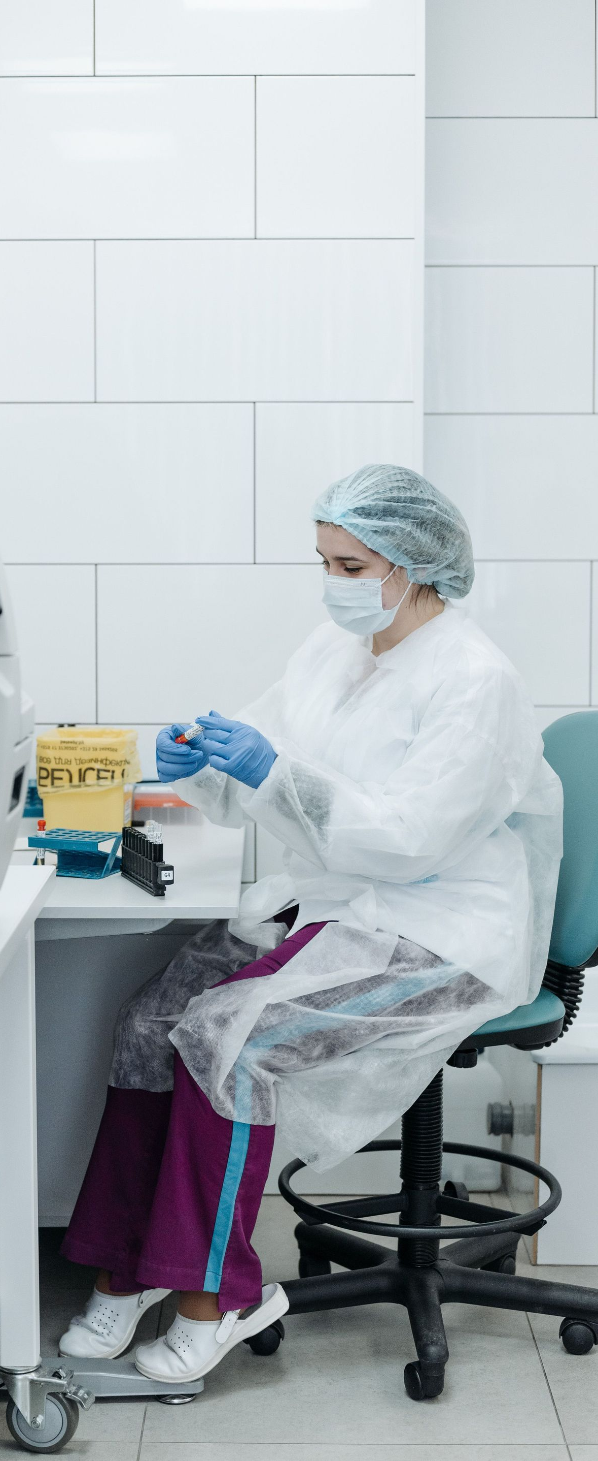 Person in white lab coat and mask working at a lab bench in a clean room with tiled walls.