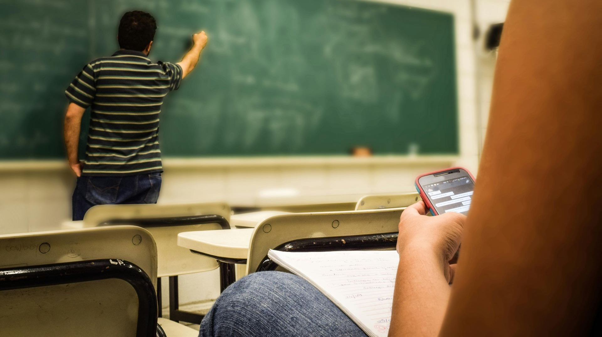 Student in classroom on phone while teacher writes on chalkboard.